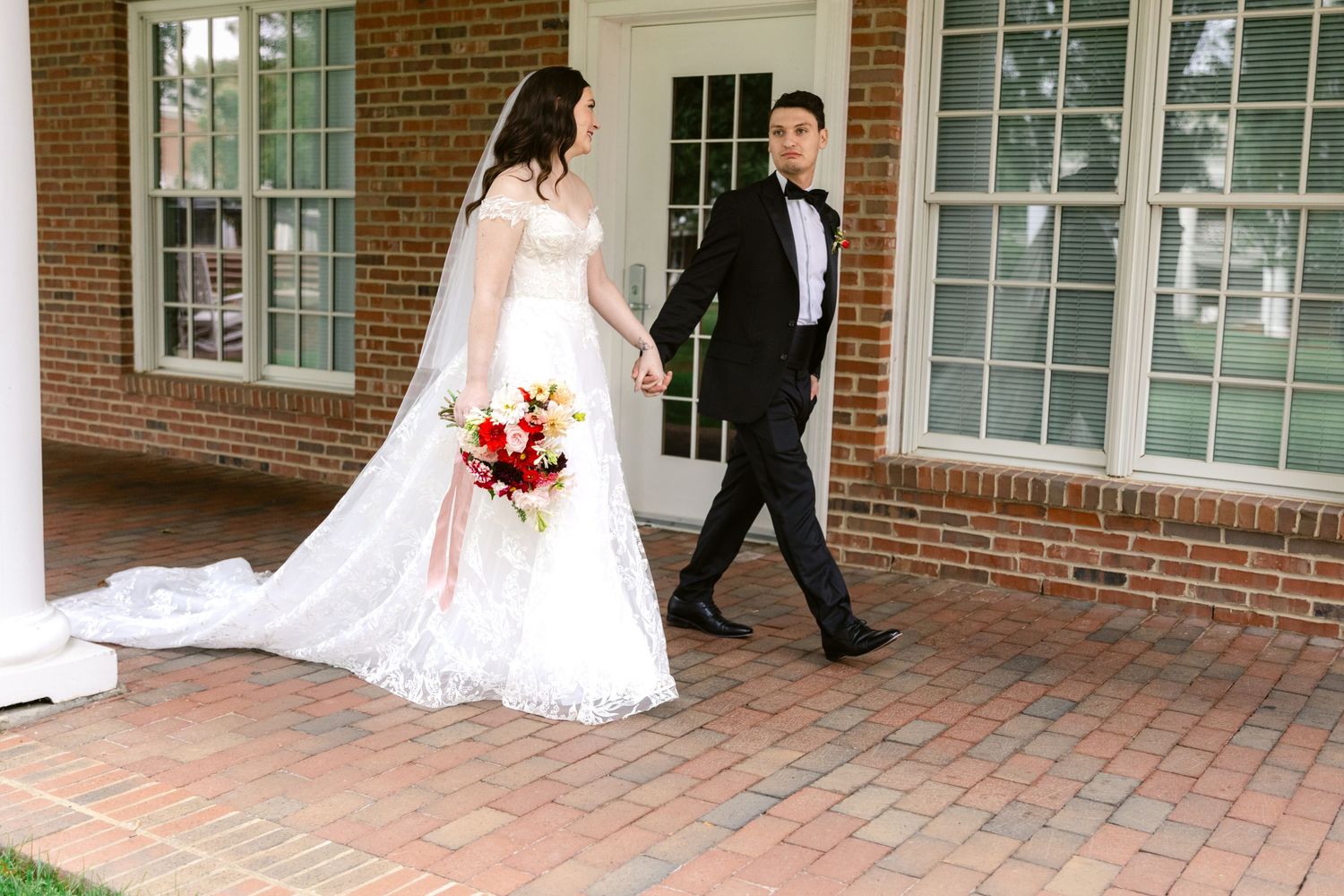 Wedding couple strolls along brick walkway outside historic brick building with white windows.