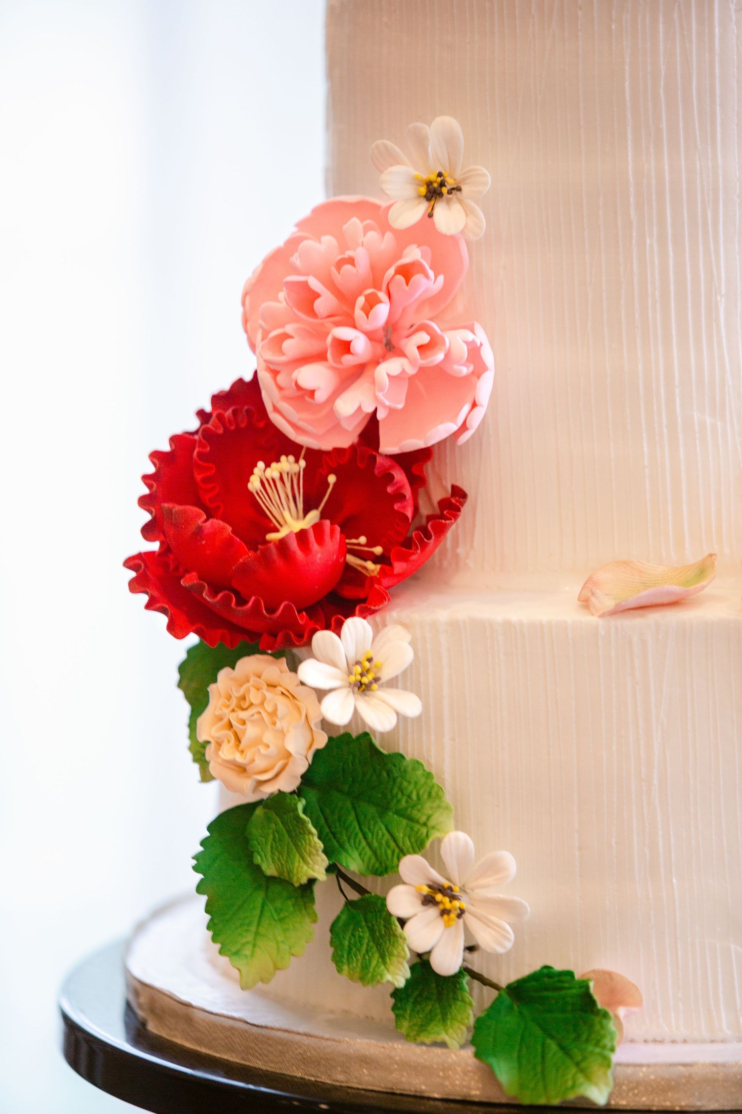 Elegant white textured wedding cake adorned with coral, red and blush sugar flowers and green foliage details.