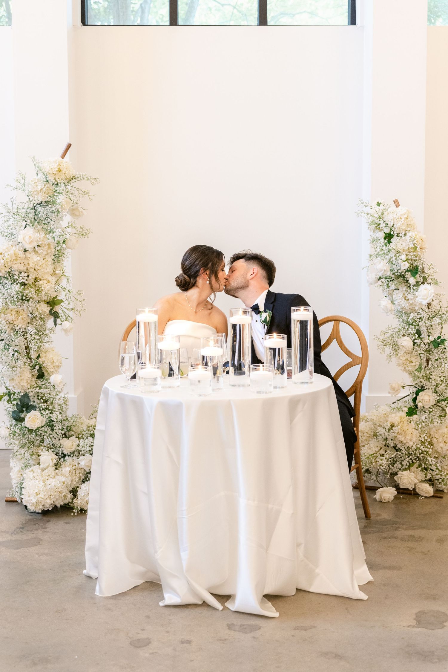 Romantic wedding couple shares an intimate moment at their sweetheart table decorated with white linens and floral arrangements.