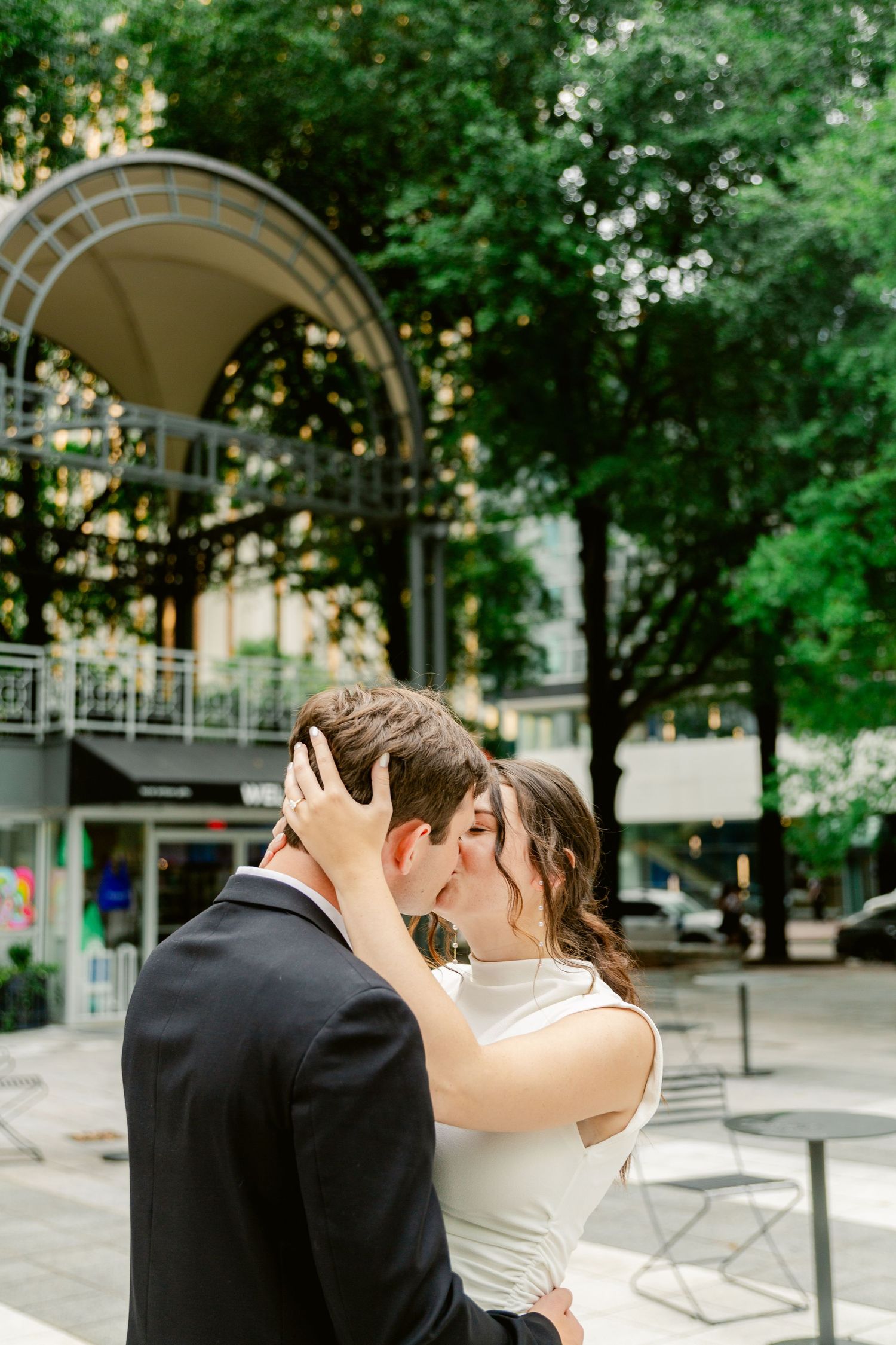 A romantic couple shares an intimate kiss in front of a city gazebo surrounded by green trees.