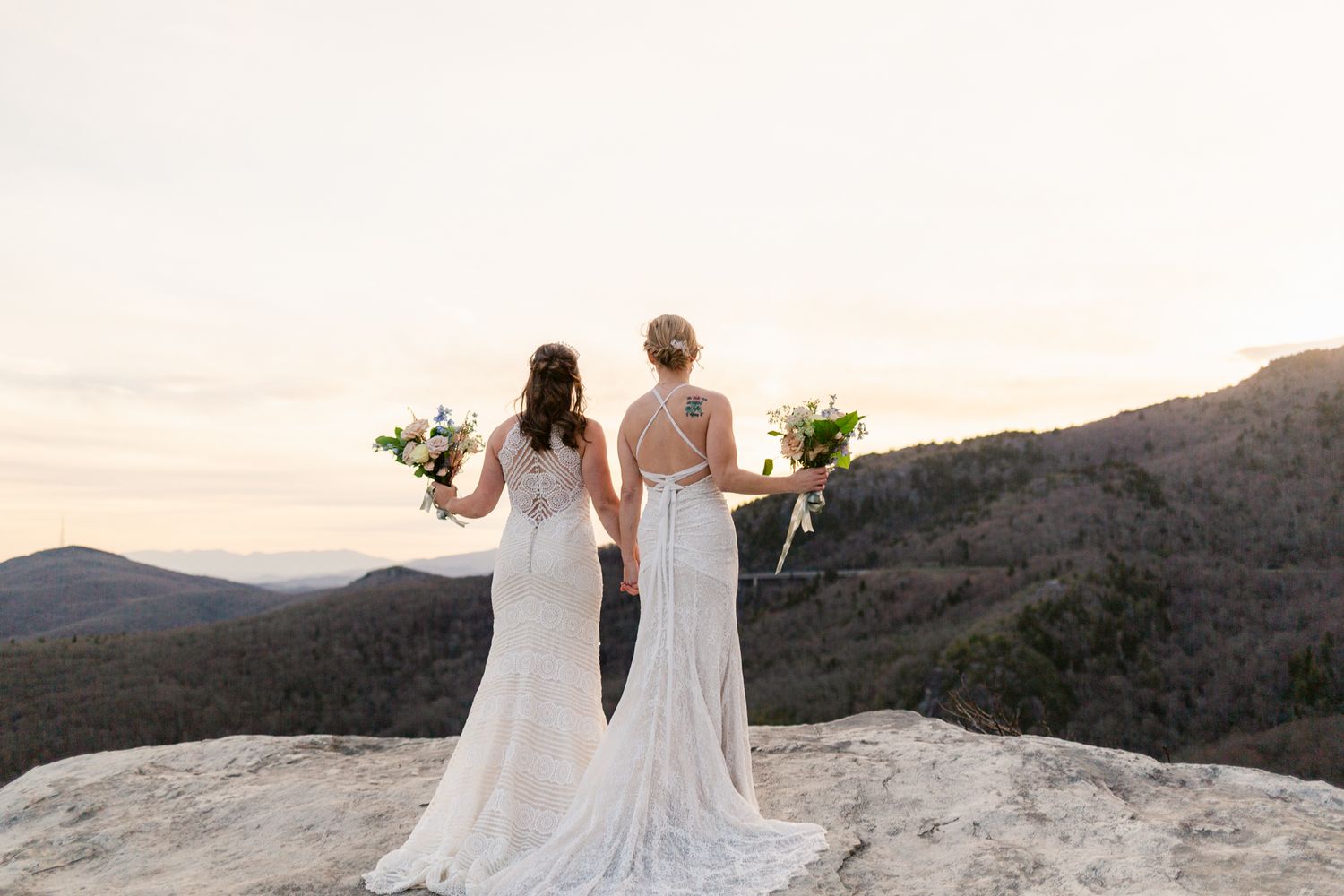 A couple in wedding attire stands on a rocky mountain overlook at sunset with scenic views in the background.