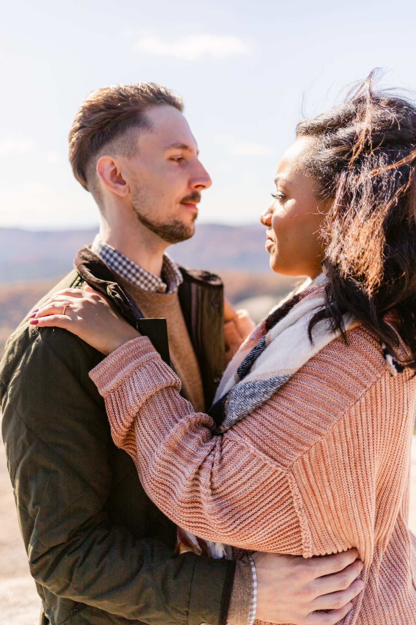 A romantic couple embraces outdoors, with one wearing a pink sweater and the other in dark clothing against a sunset backdrop.
