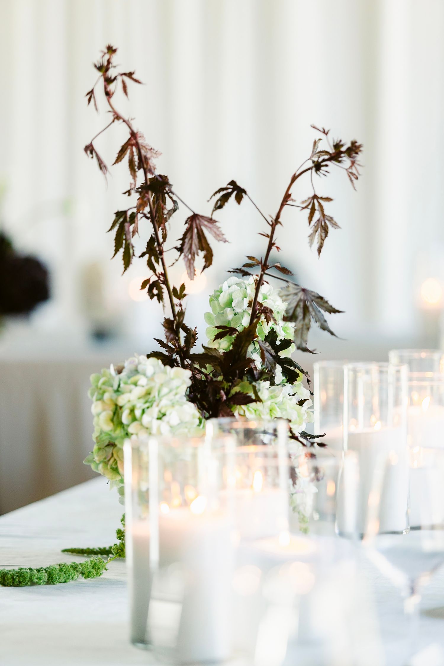 Elegant table setting with dark branches, white hydrangeas and glowing candles in glass holders on a white tablecloth.