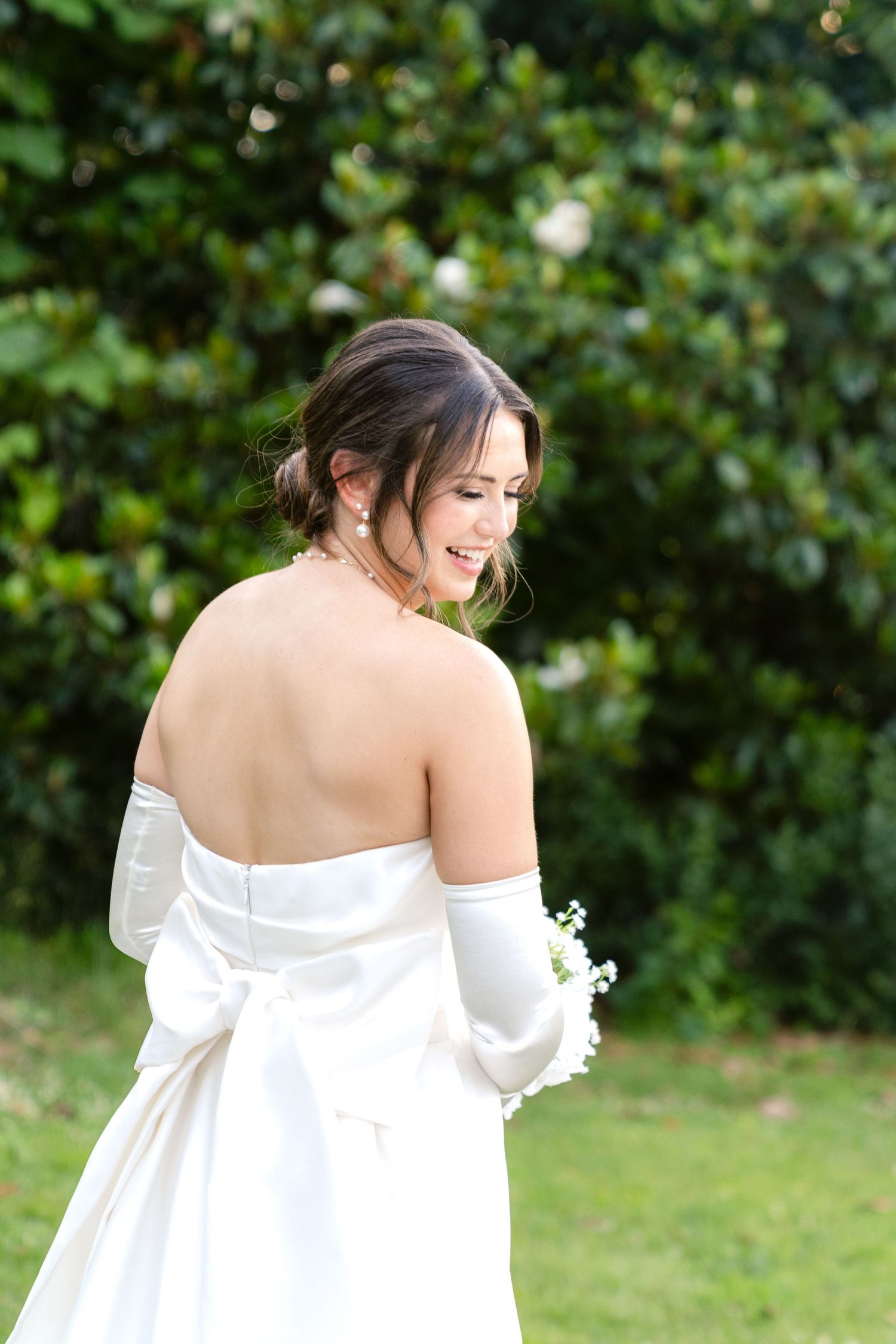 A bride in an elegant off-shoulder white wedding dress stands in a garden setting with green foliage in the background.