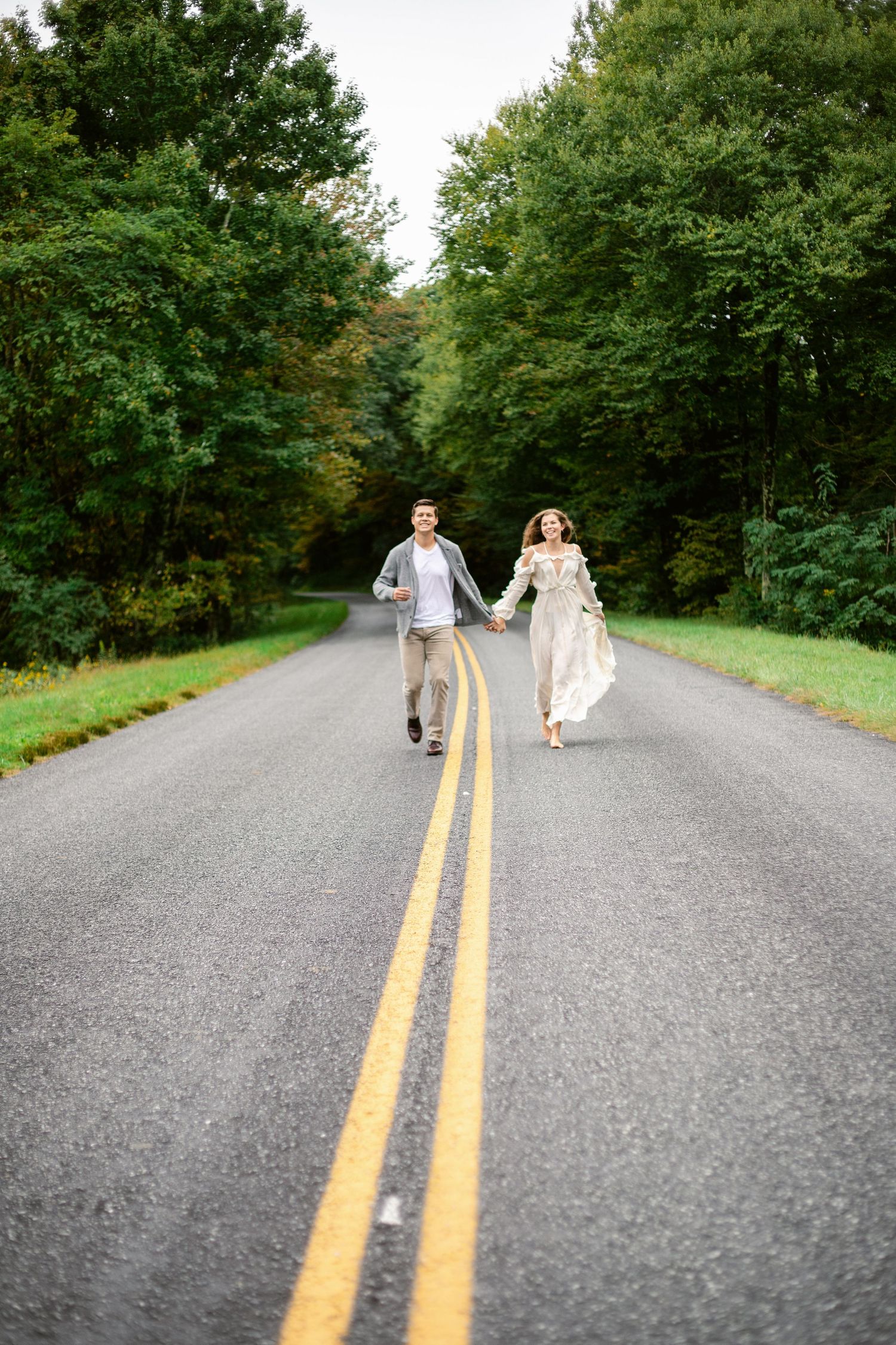 Couple in wedding attire running playfully down a road surrounded by lush green trees.