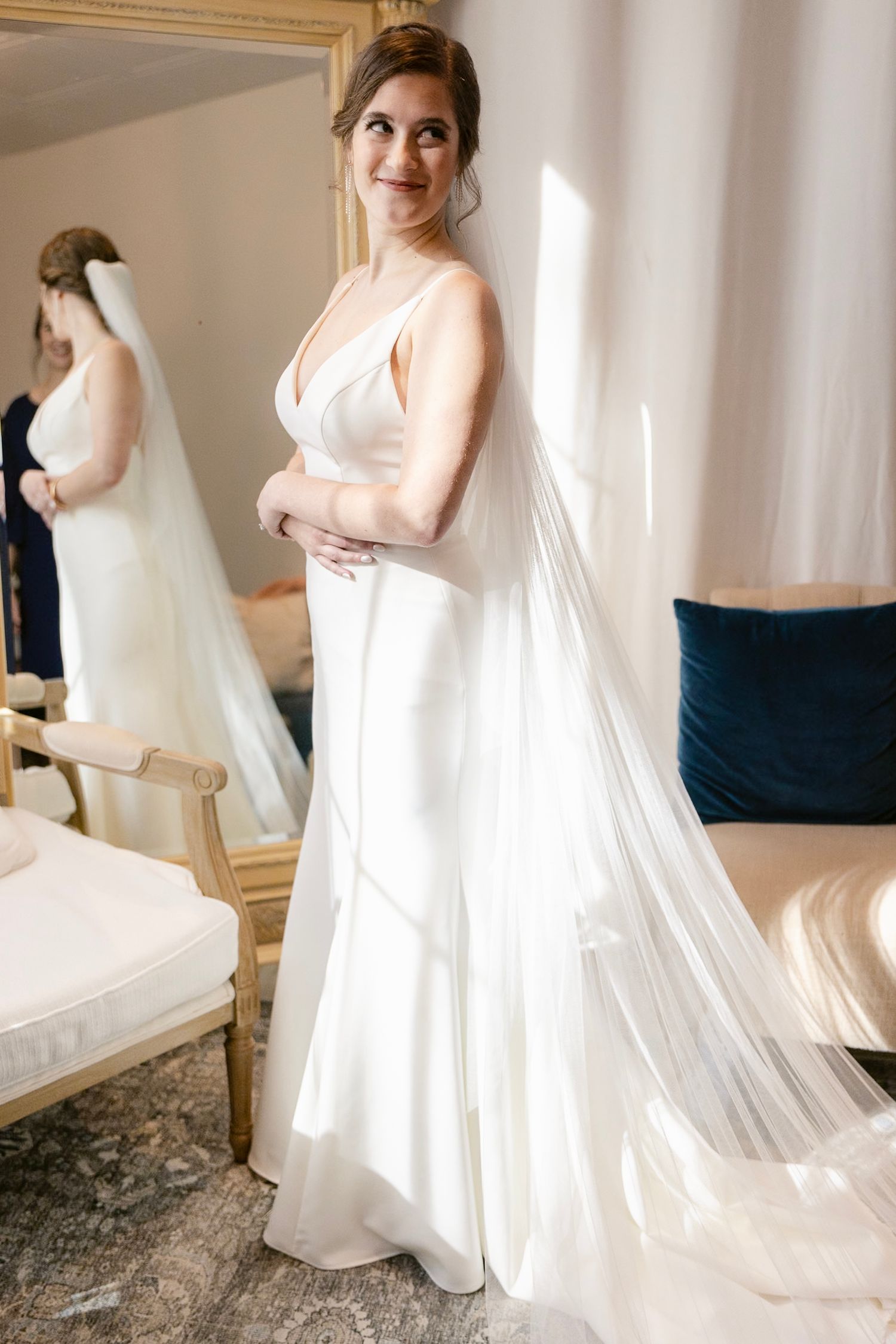 A bride in an elegant white satin gown with a flowing veil stands in a sunlit room next to a mirror.