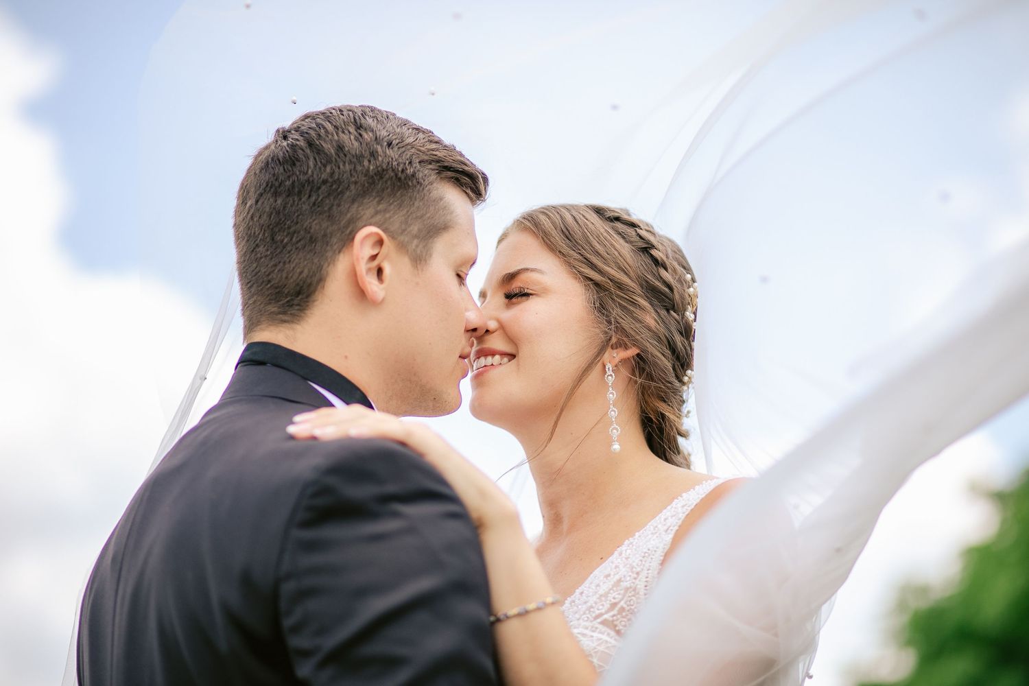 Joyful wedding portrait of couple kissing with flowing veil against blue sky.