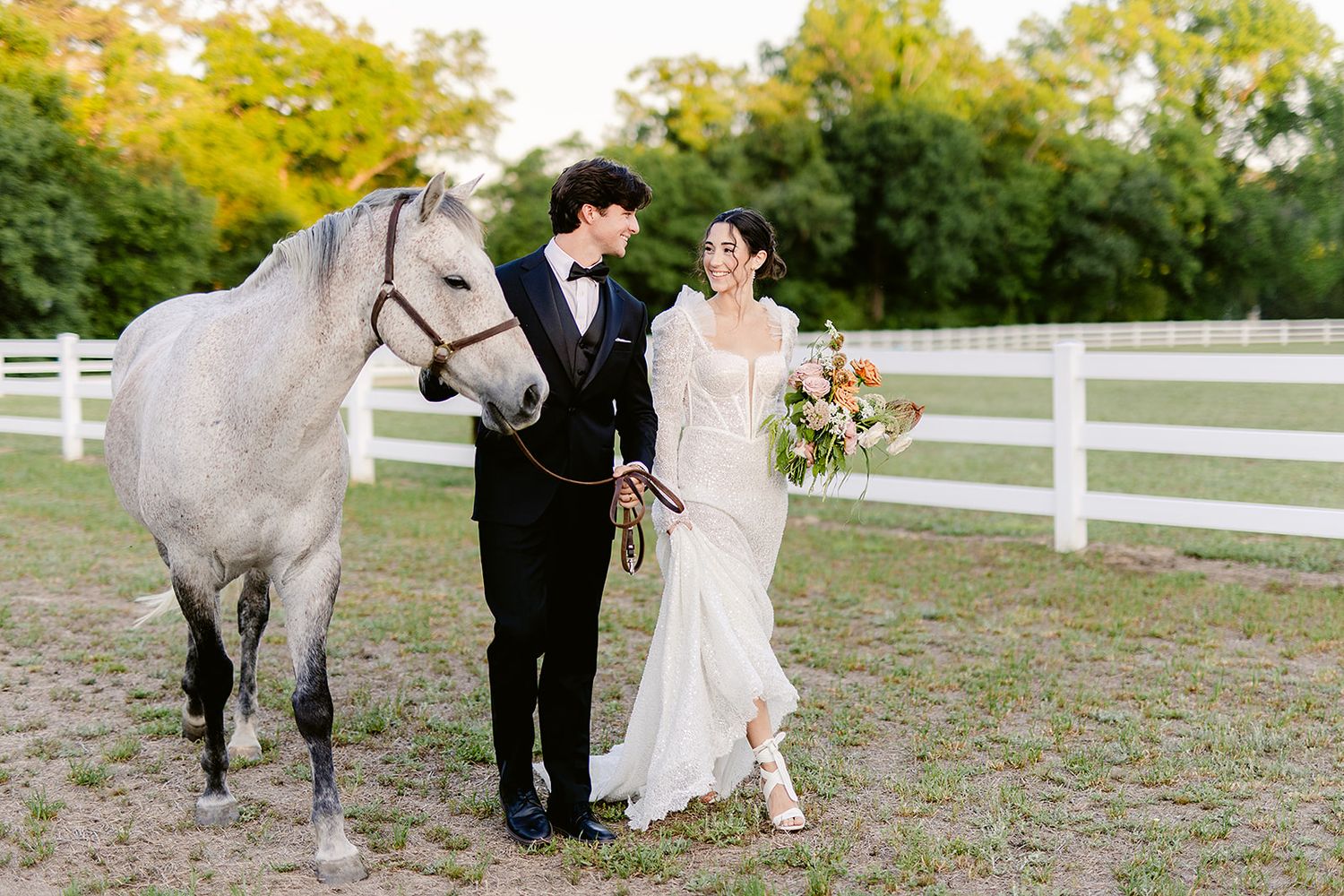 A beautiful white horse stands beside a couple in formal attire at a rustic farm with white fencing.