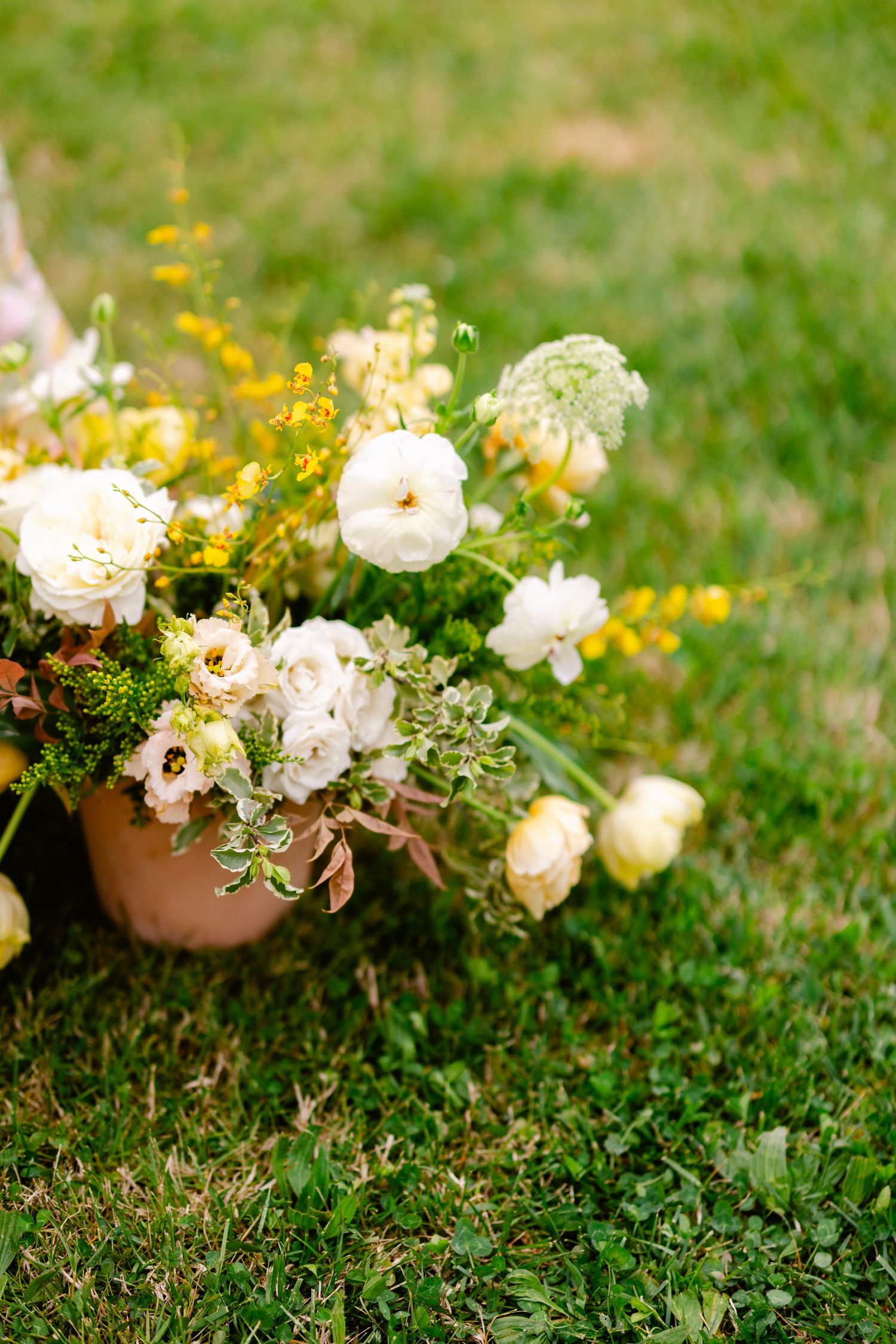 White floral arrangements in terracotta pots rest on green grass with yellow wildflowers in the background.