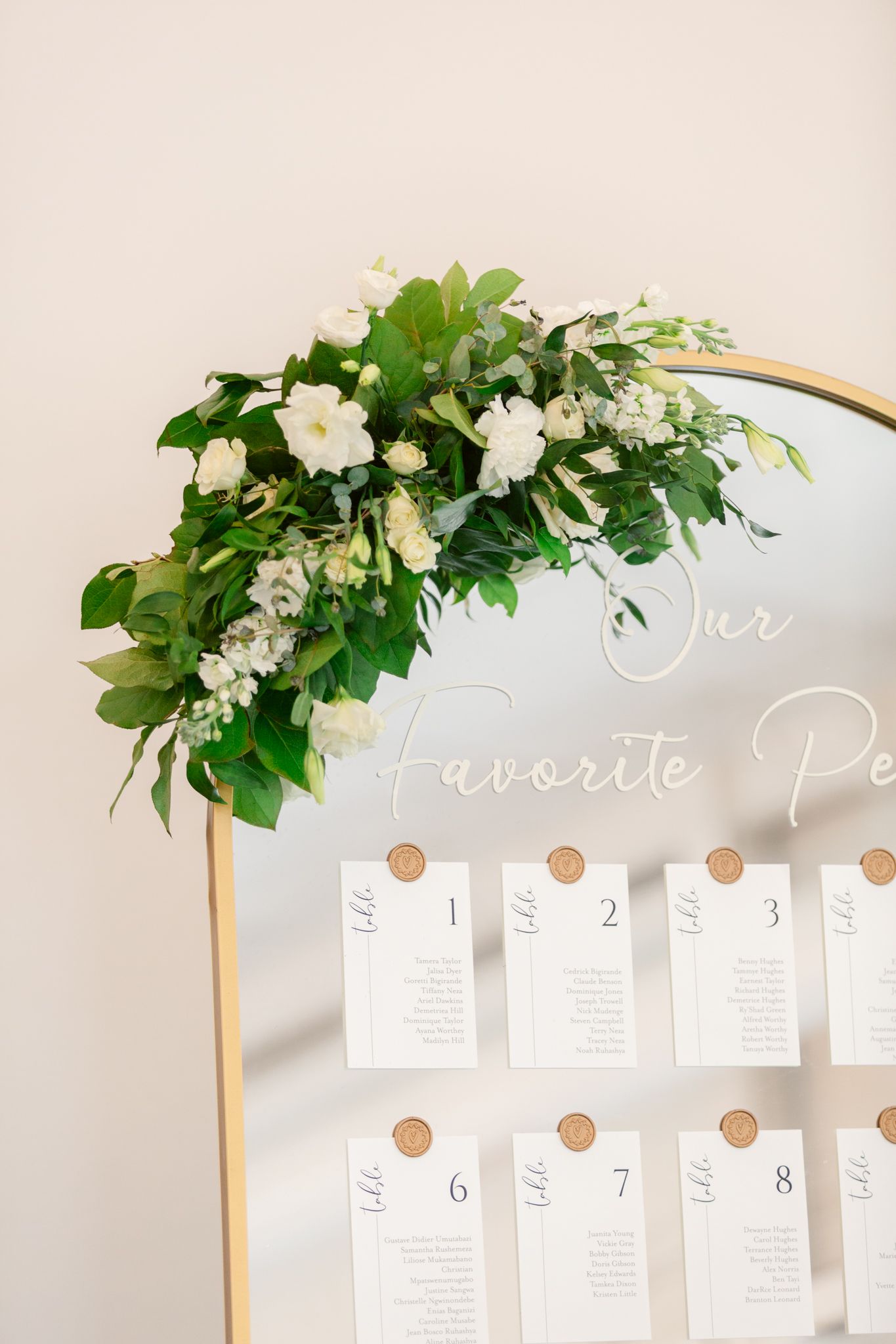 Wedding seating chart display with greenery and white floral arch decoration against white wall.