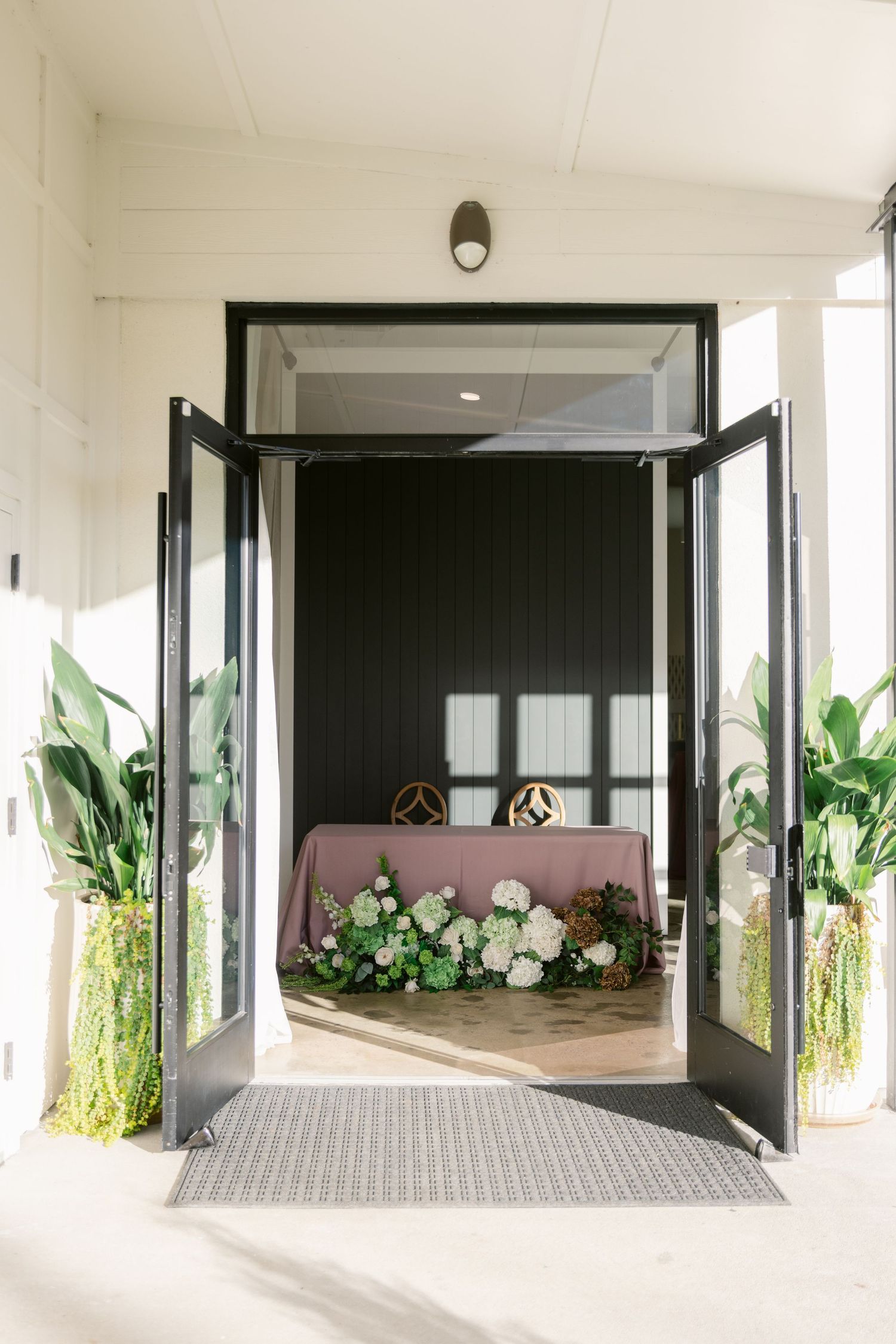 Modern building entrance with dark framed glass doors and decorative floral arrangements in white and pink tones.