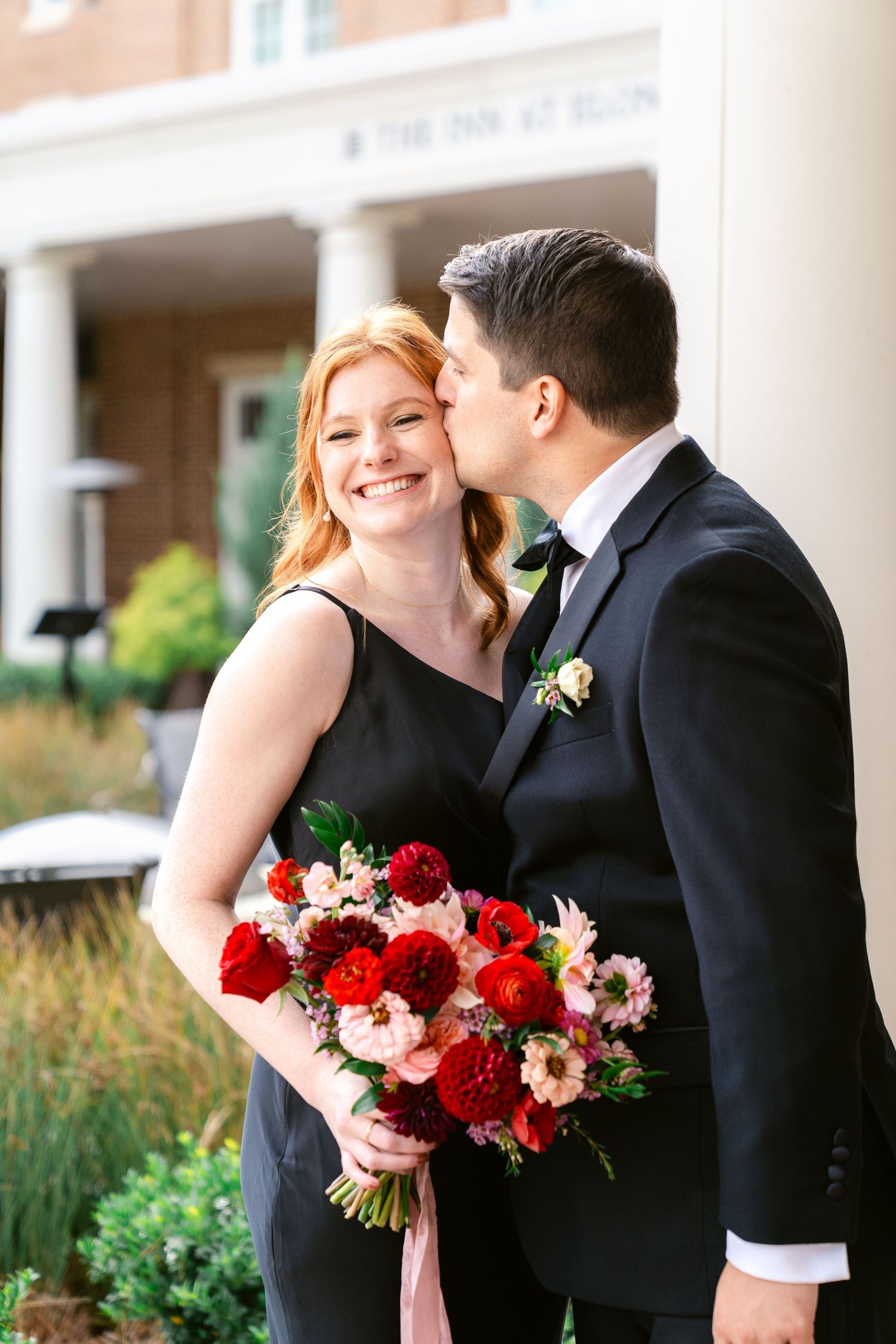 Romantic wedding couple shares tender moments outdoors with vibrant red and pink floral bouquet.