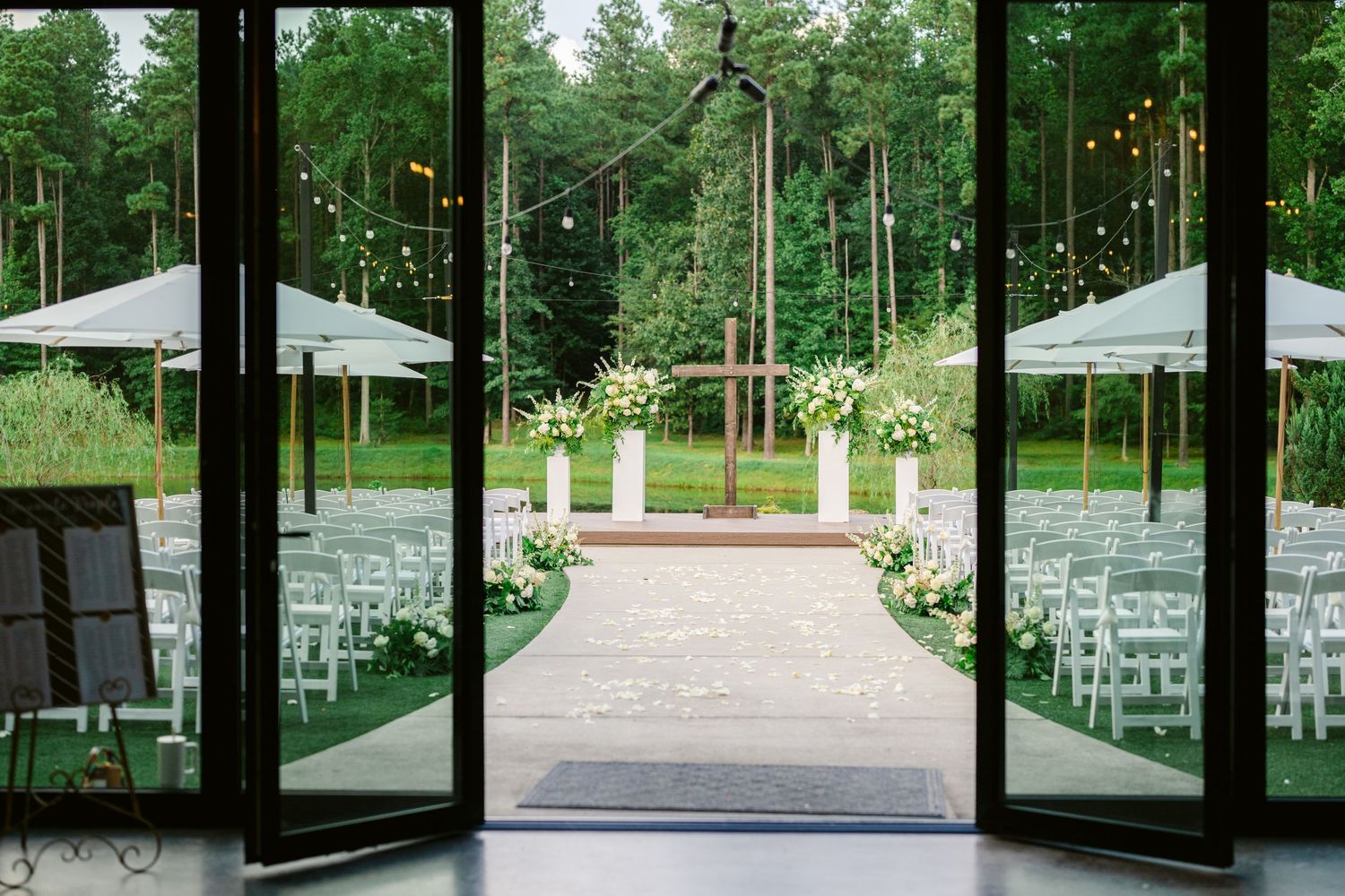 White chairs and floral arrangements line a garden path for an outdoor wedding ceremony in a serene forest setting.