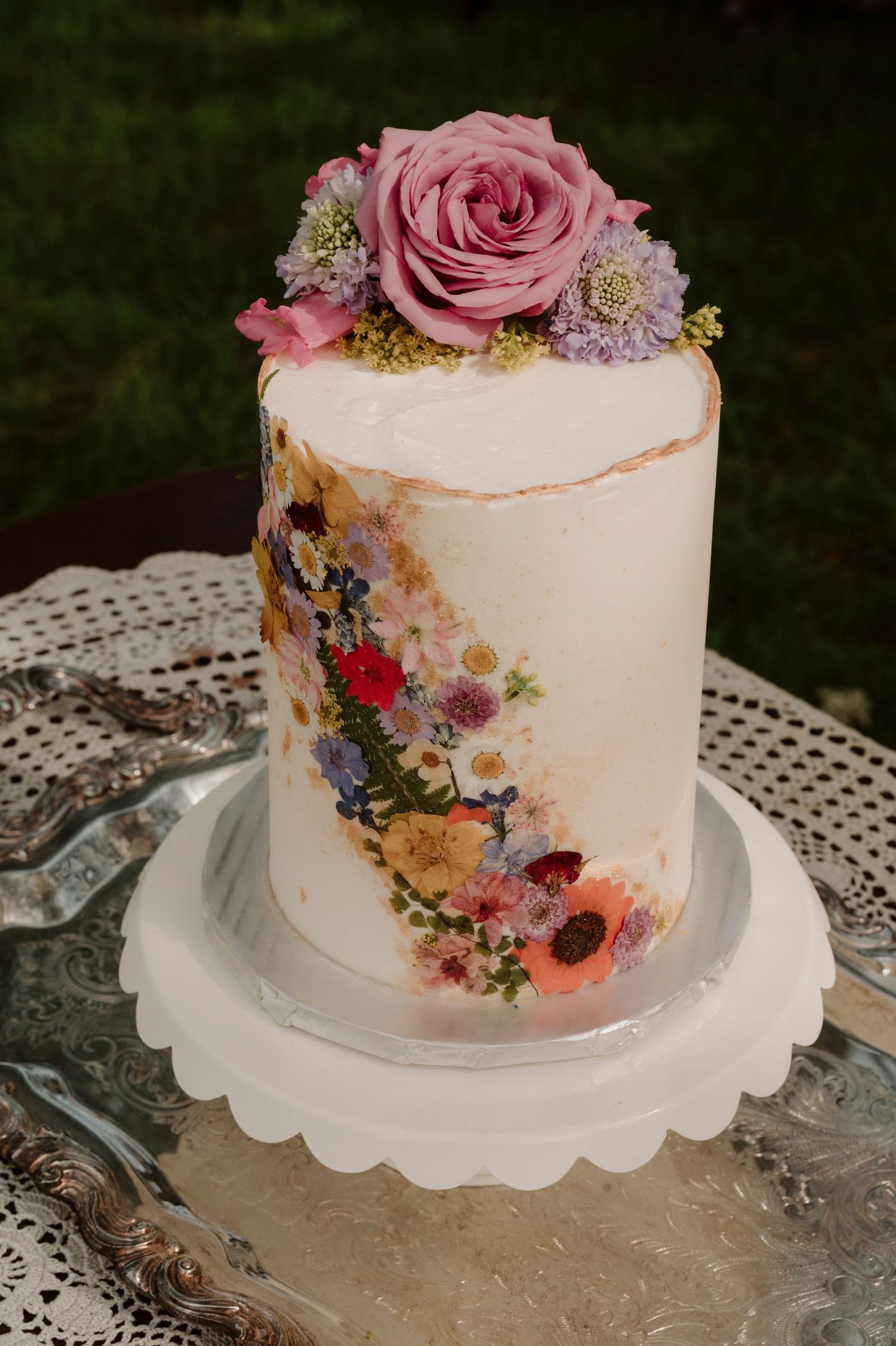 A single-tier white wedding cake decorated with pressed flowers and topped with a fresh pink rose.