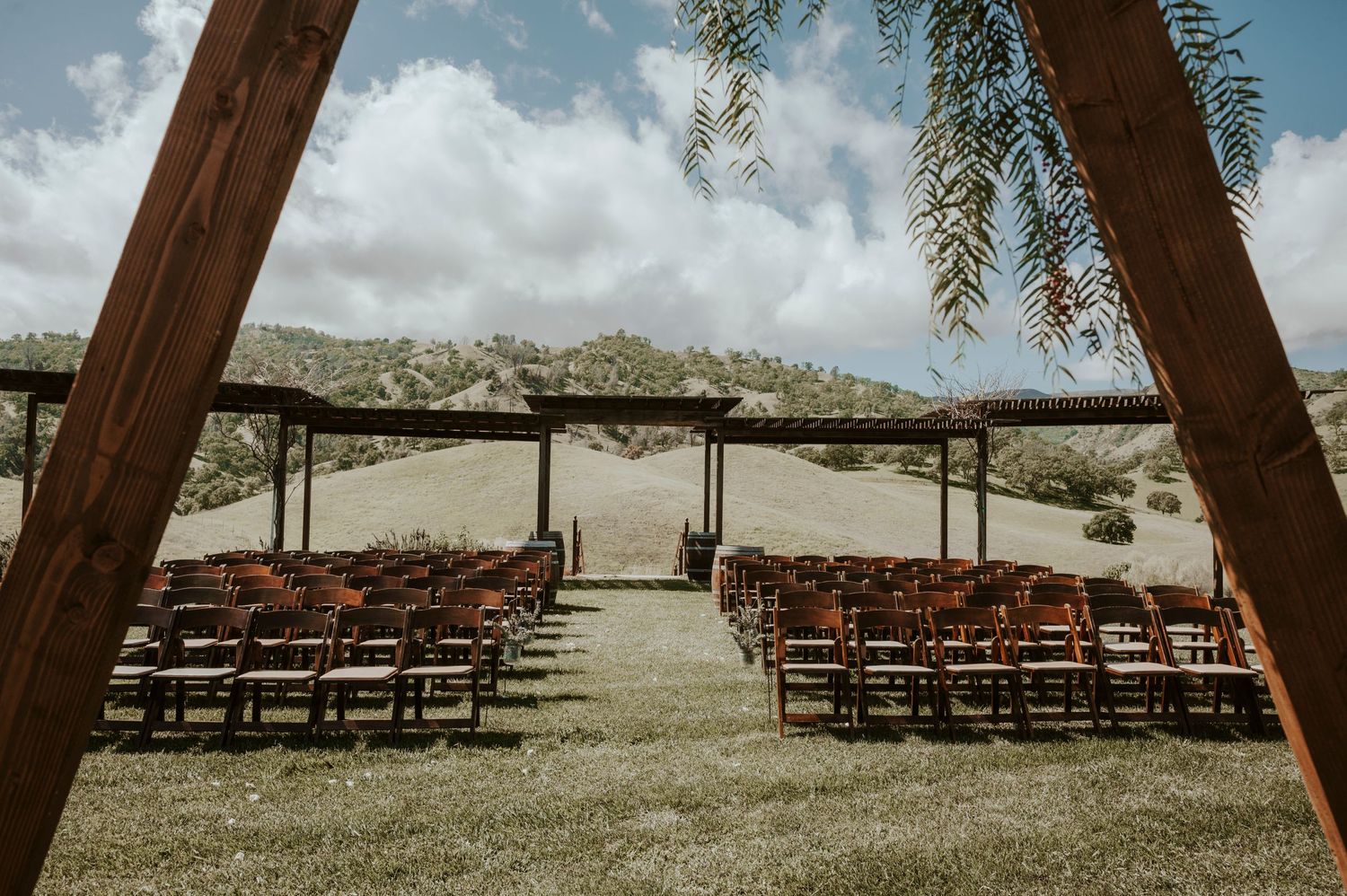 Wooden chairs arranged for an outdoor wedding ceremony under a rustic pergola against a mountain backdrop.