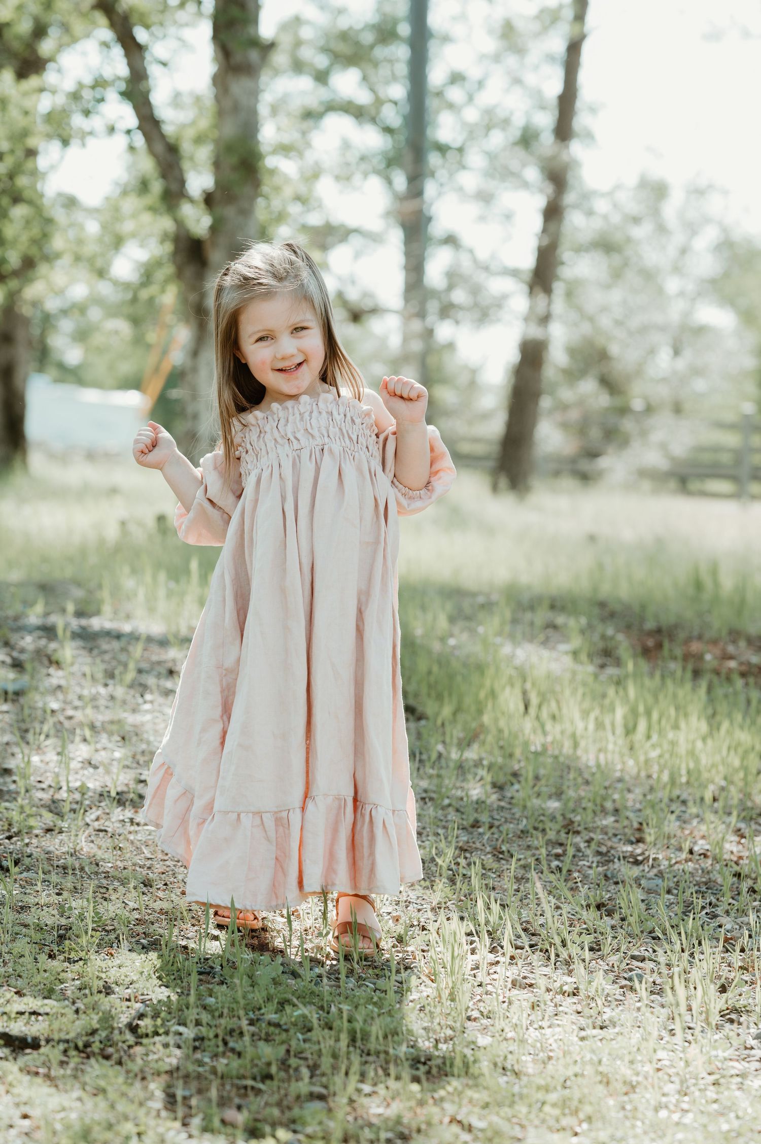 A soft pink tiered ruffle dress worn in a serene outdoor setting with sunlit grass and trees.