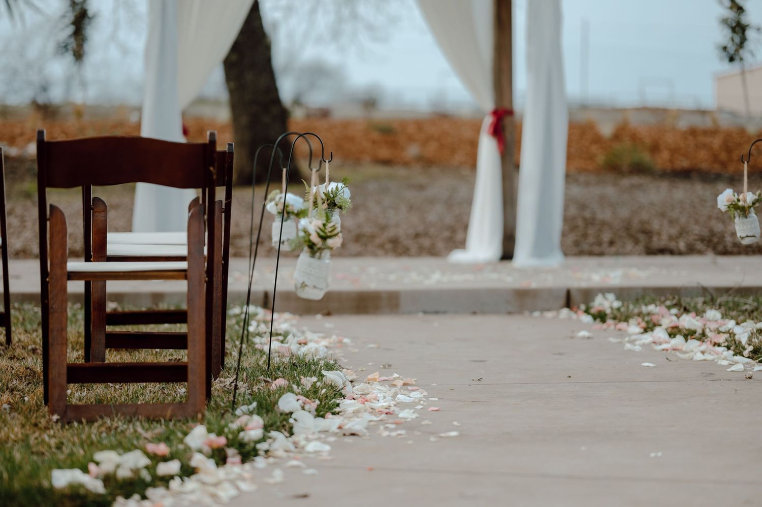 Wooden chairs and scattered petals line an outdoor wedding aisle with white draping.