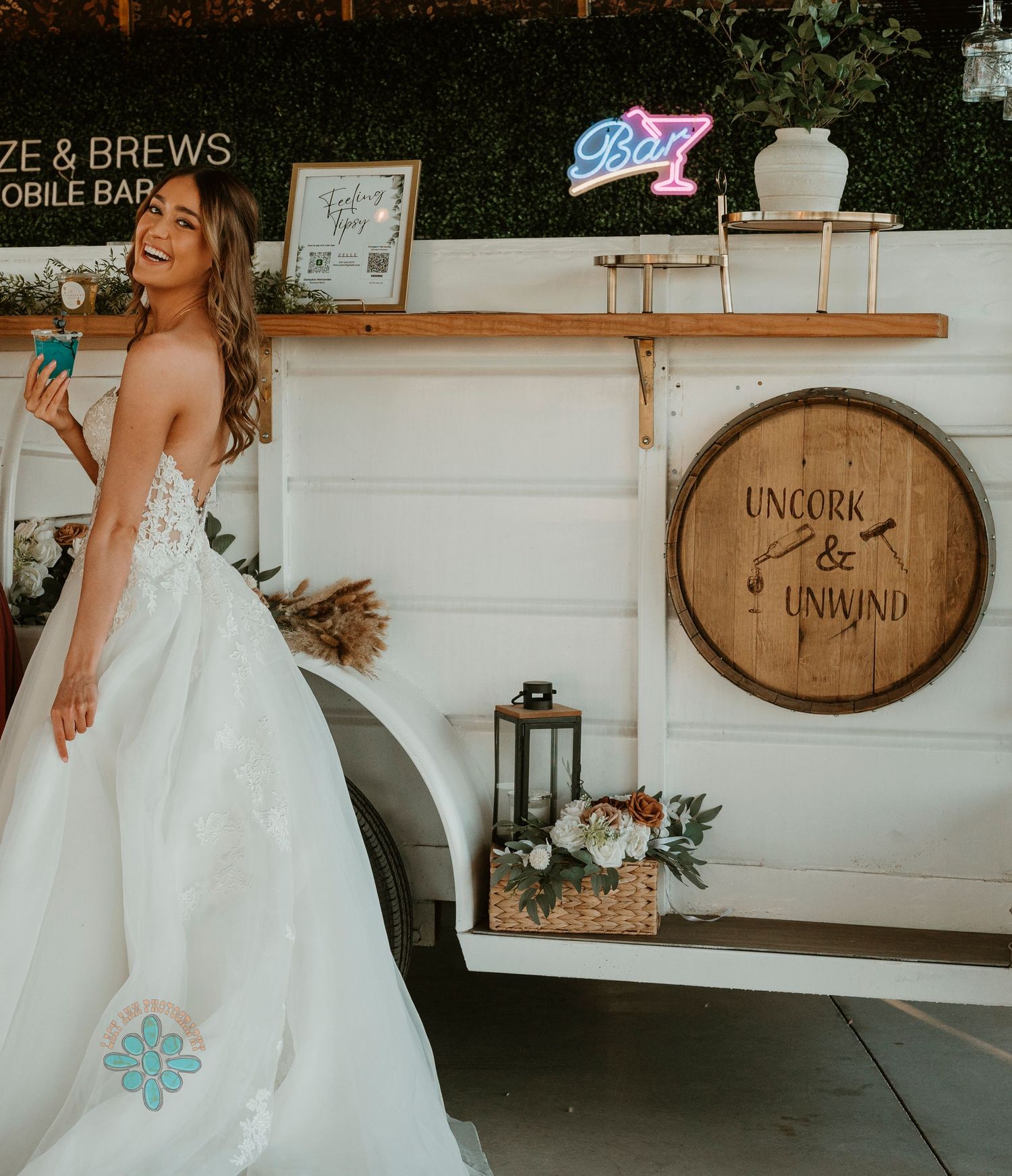 Mobile bar setup with greenery wall and wine barrel decor at wedding venue.