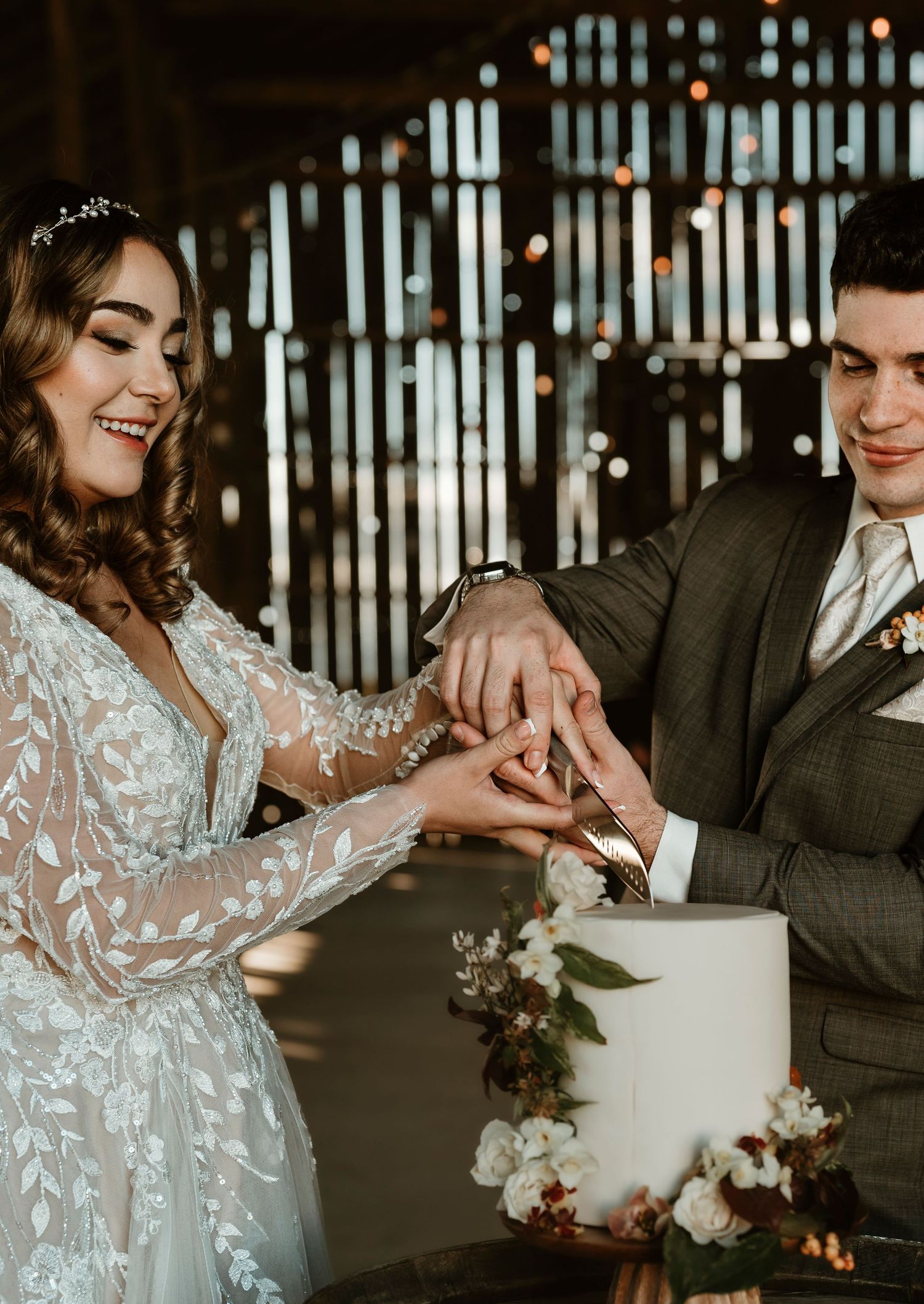 Newlyweds cutting their elegant white wedding cake together in a rustic venue with string lights.