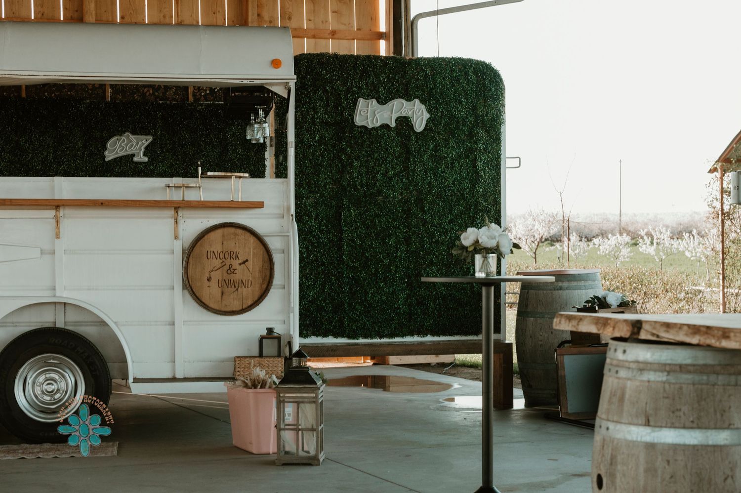 Rustic outdoor venue with vintage white bus, wooden barrels, and artificial grass wall decor.