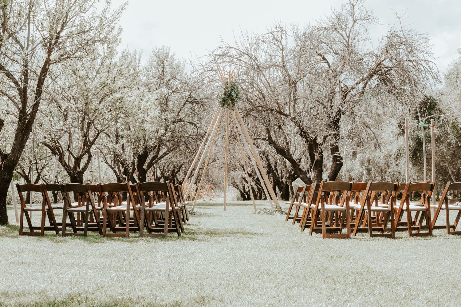 Wooden folding chairs arranged in rows for an outdoor wedding ceremony with a triangle arch centerpiece.