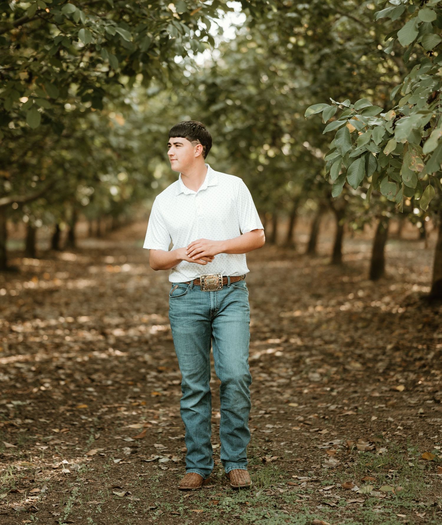 Young man in white polo and jeans standing in a tree orchard, looking up thoughtfully.