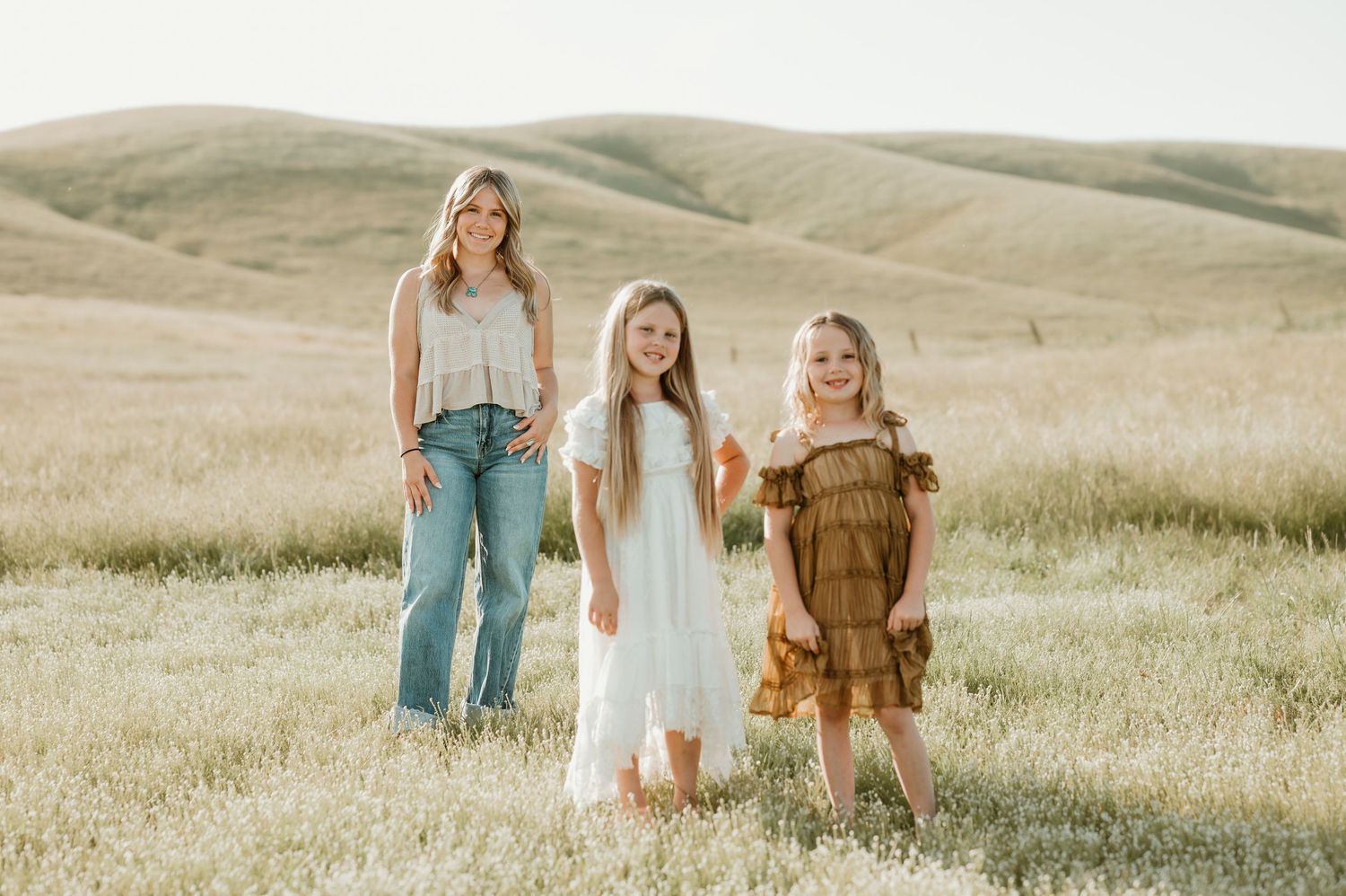 Three people in casual clothing stand together on a grassy hillside during golden hour.