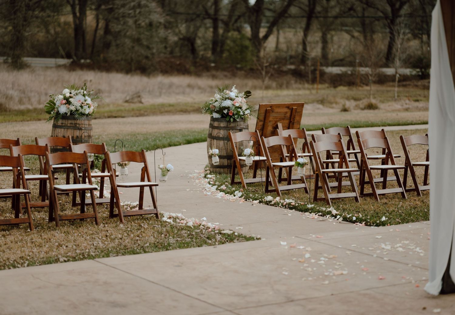 Rustic outdoor wedding ceremony setup with wooden chairs arranged along a curved path and white floral arrangements.