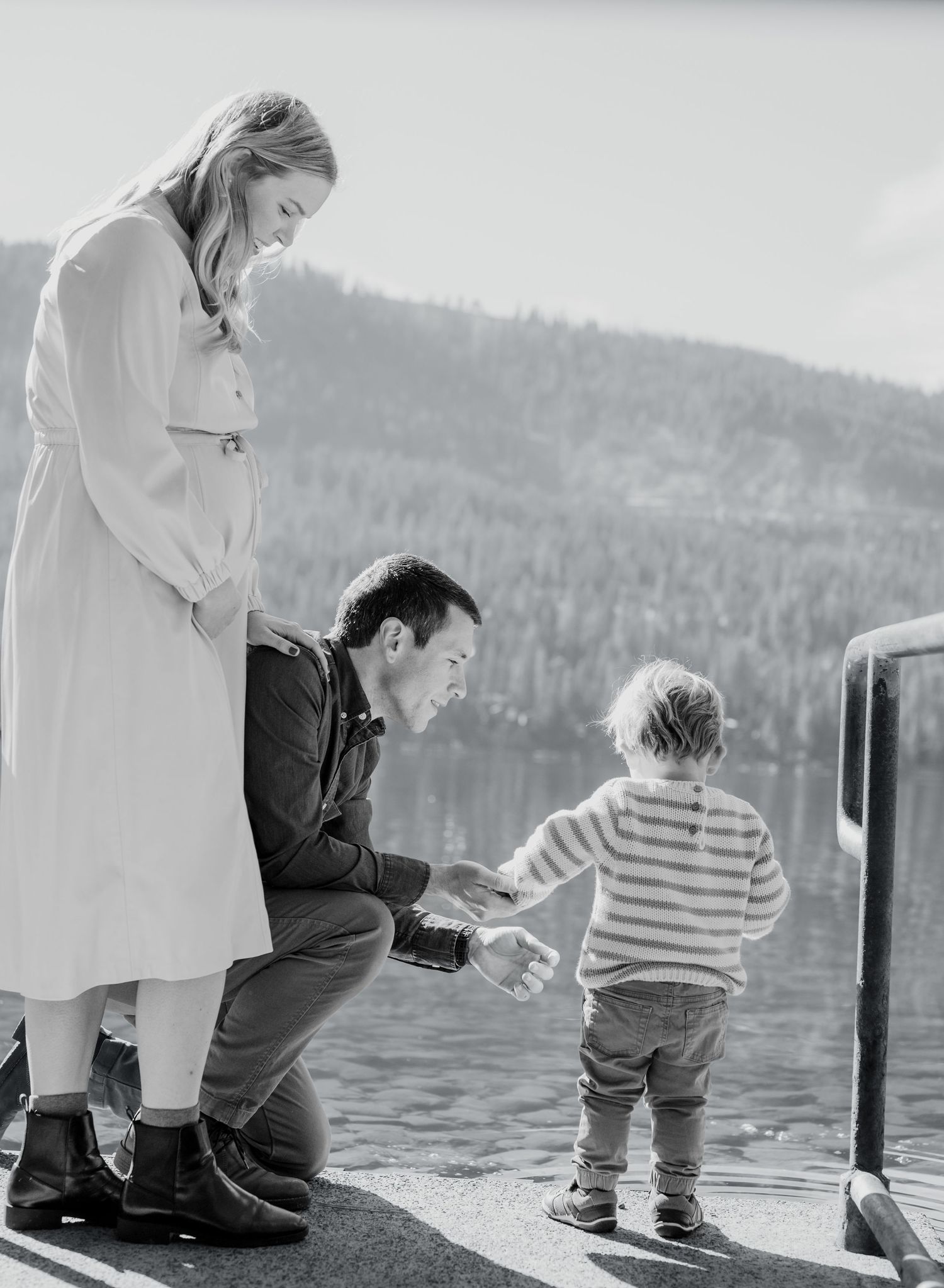 Family enjoying a quiet moment together on a dock by a lake with mountains in the background, captured in black and white.