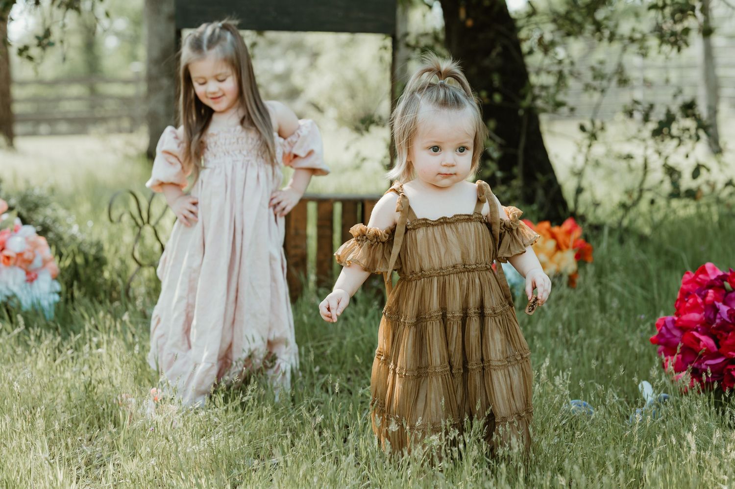 Children wearing vintage-style dresses play together in a sunlit garden with vibrant pink flowers.