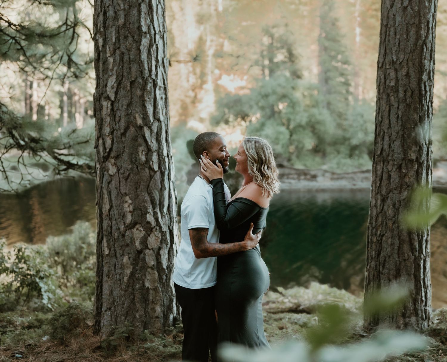 A romantic couple embraces between tall pine trees near a tranquil lake during golden hour.