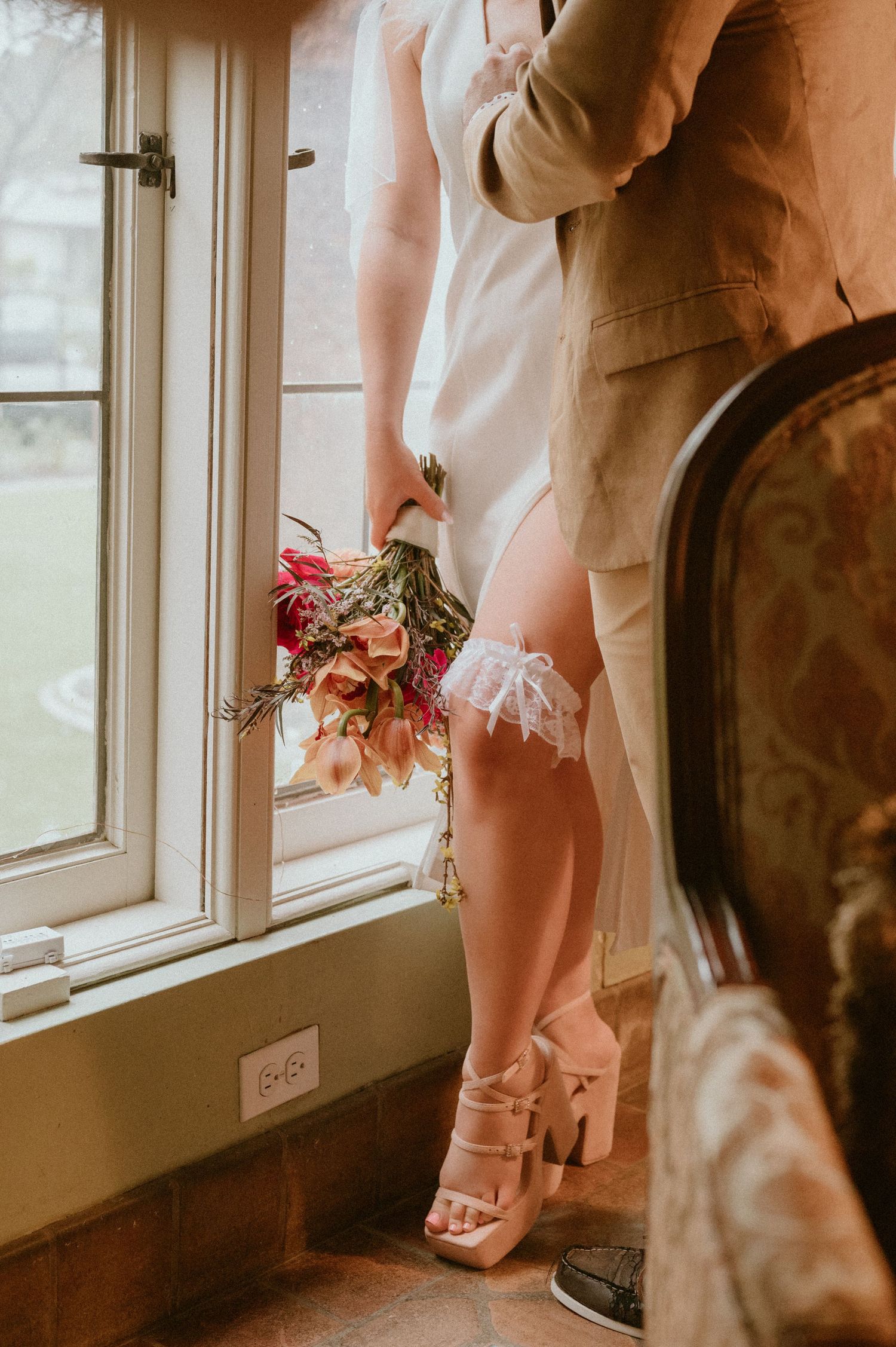 Close-up view of white wedding shoes and pink floral bouquet details by a sunlit window.