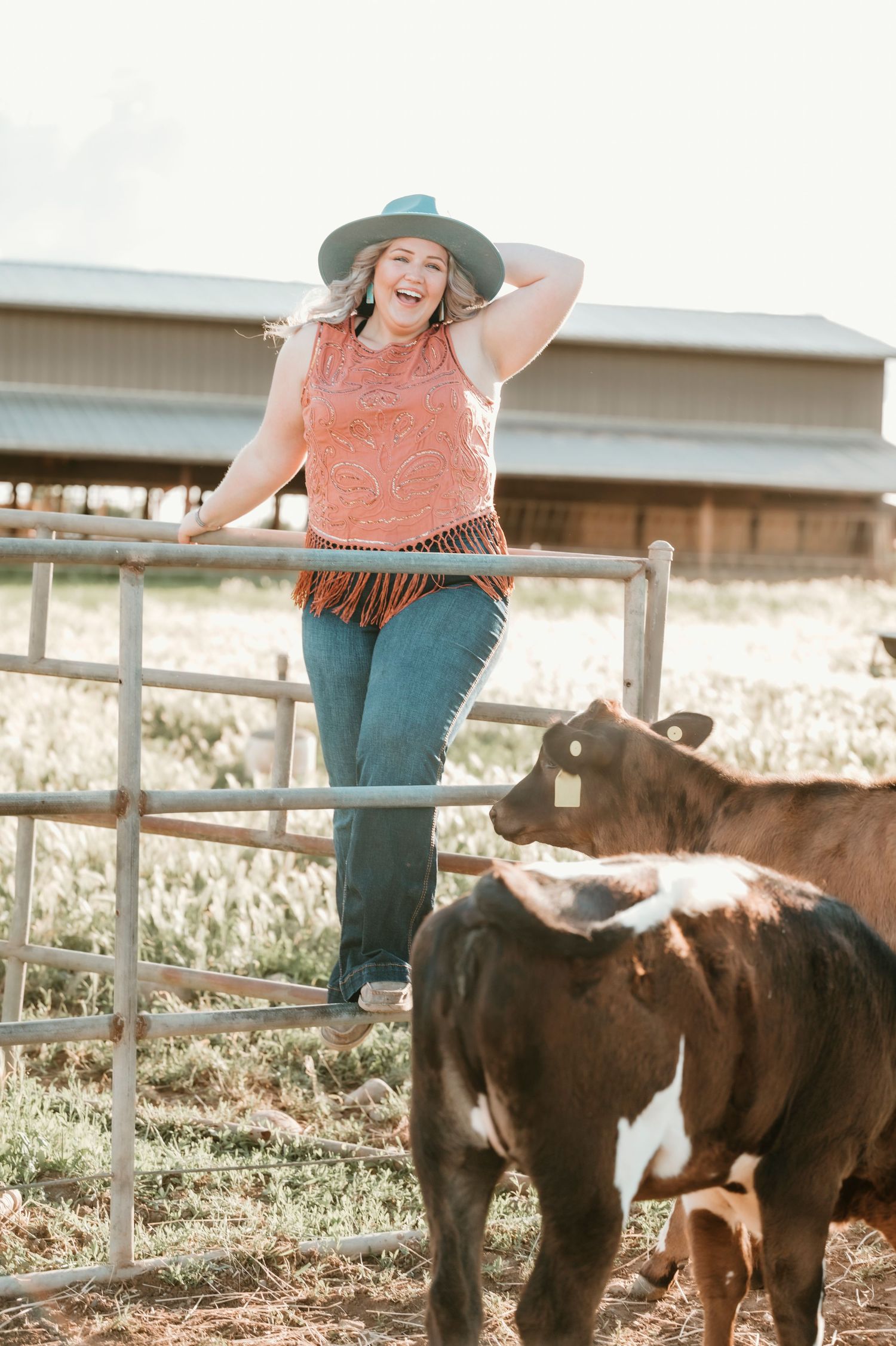 A person in western wear leans on a fence near cattle at a ranch during sunset.