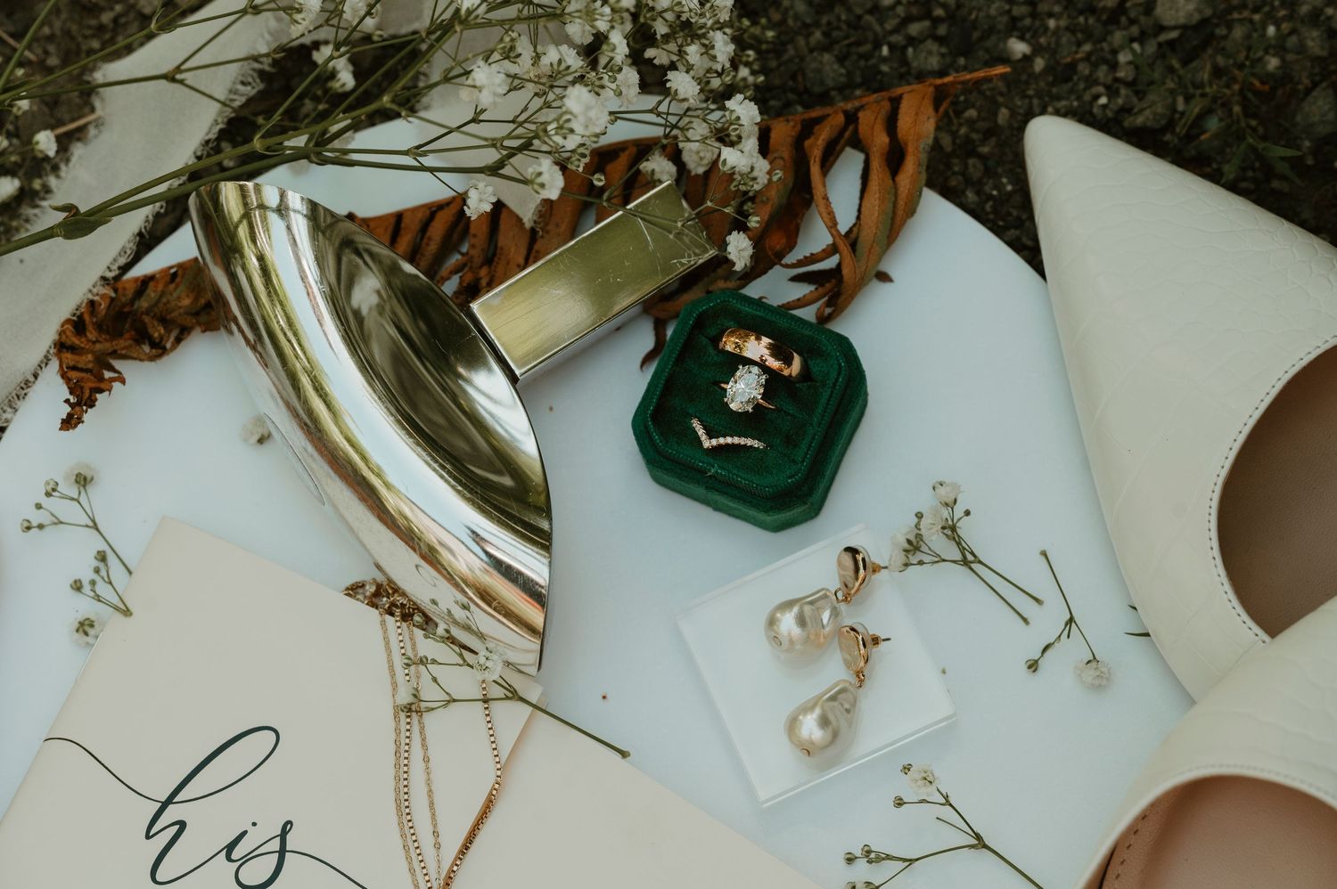Wedding details flatlay with perfume bottle, jewelry, and delicate floral elements on white surface.