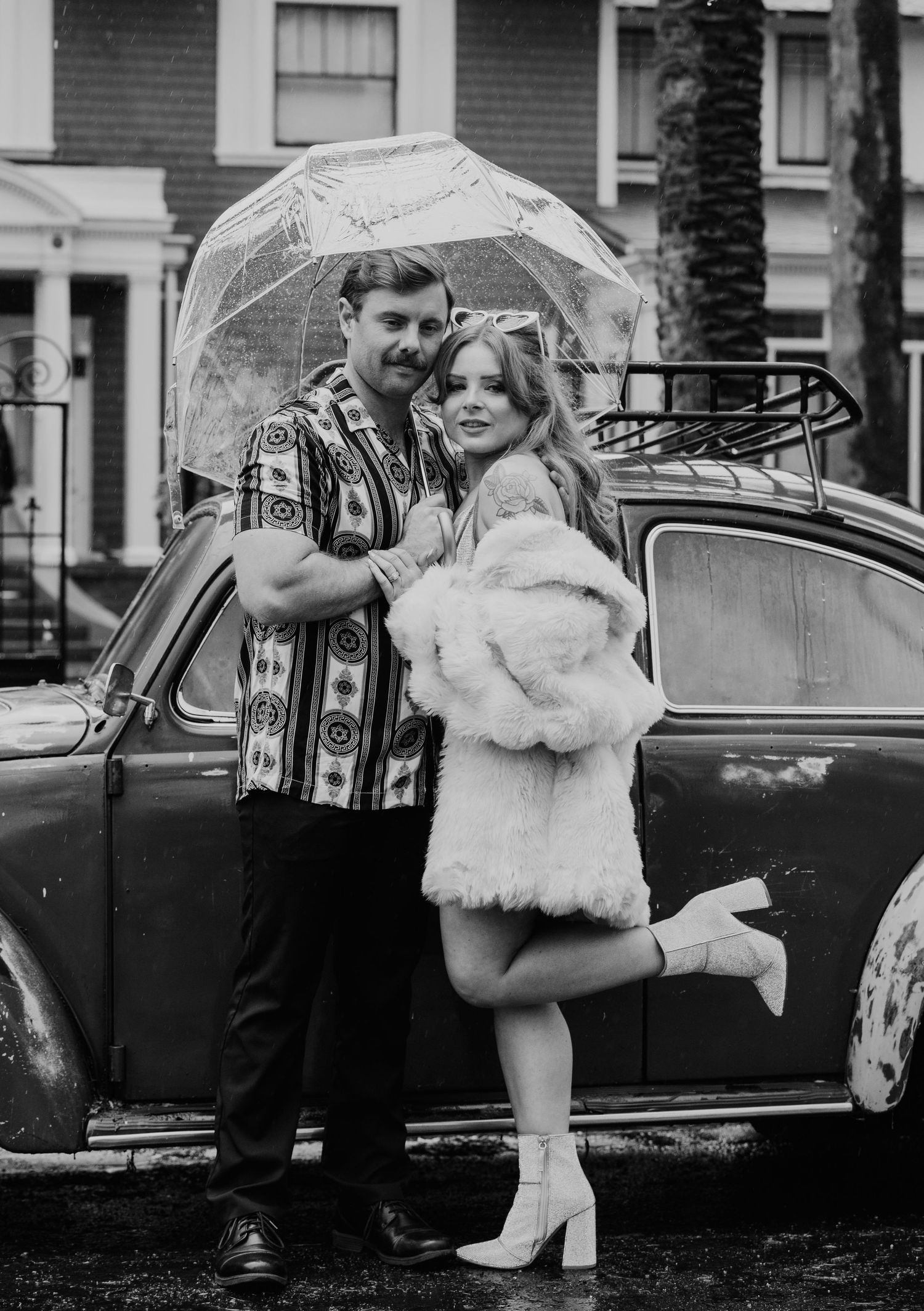 A couple poses with a clear umbrella next to a vintage car, with the woman wearing white boots and a fur coat.