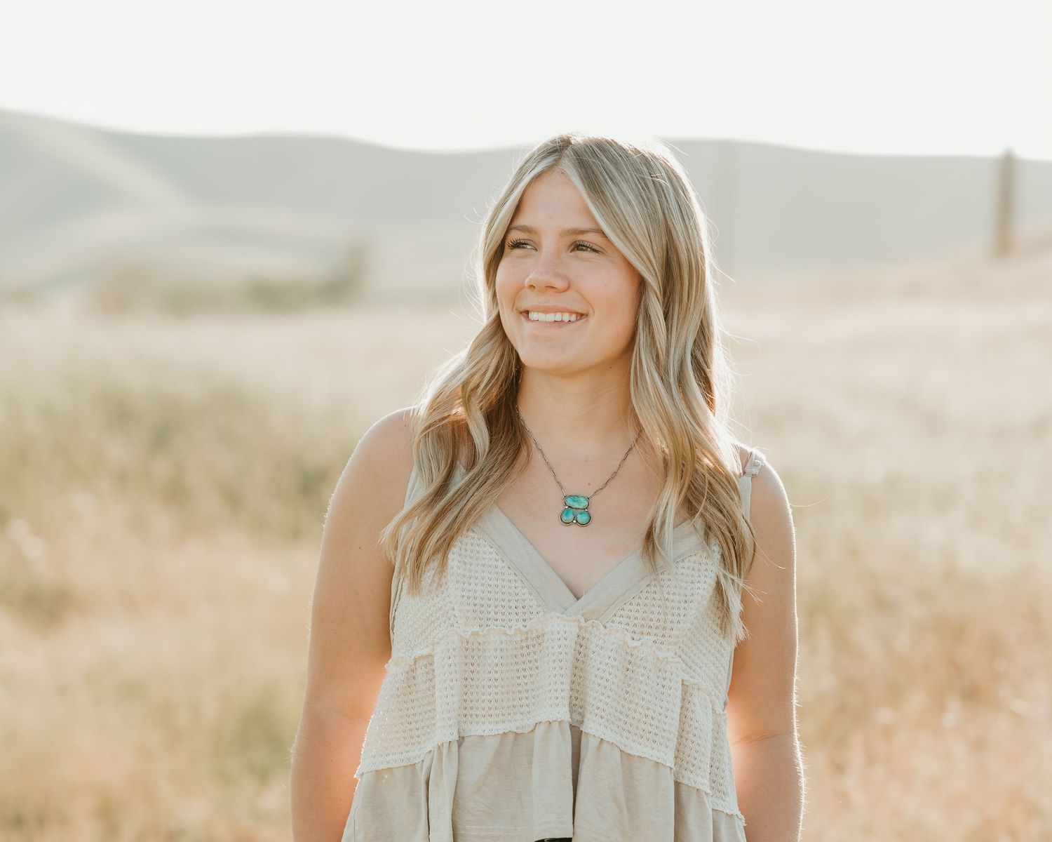 Person with long blonde hair wearing beige outfit stands in golden sunlit field at sunset.