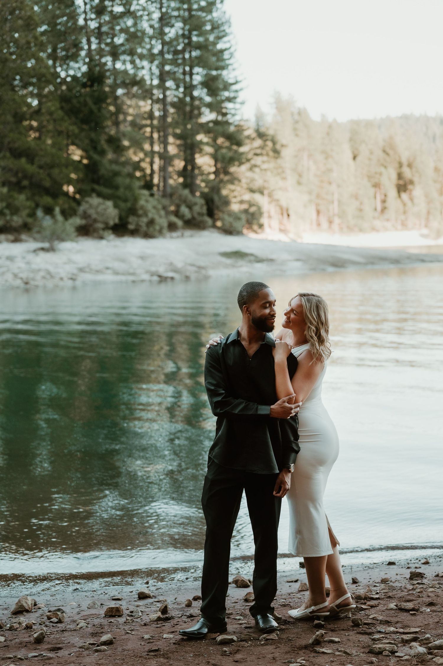 A winter white dress contrasts against evergreens while a couple stands along a lake shore in an atmospheric setting.