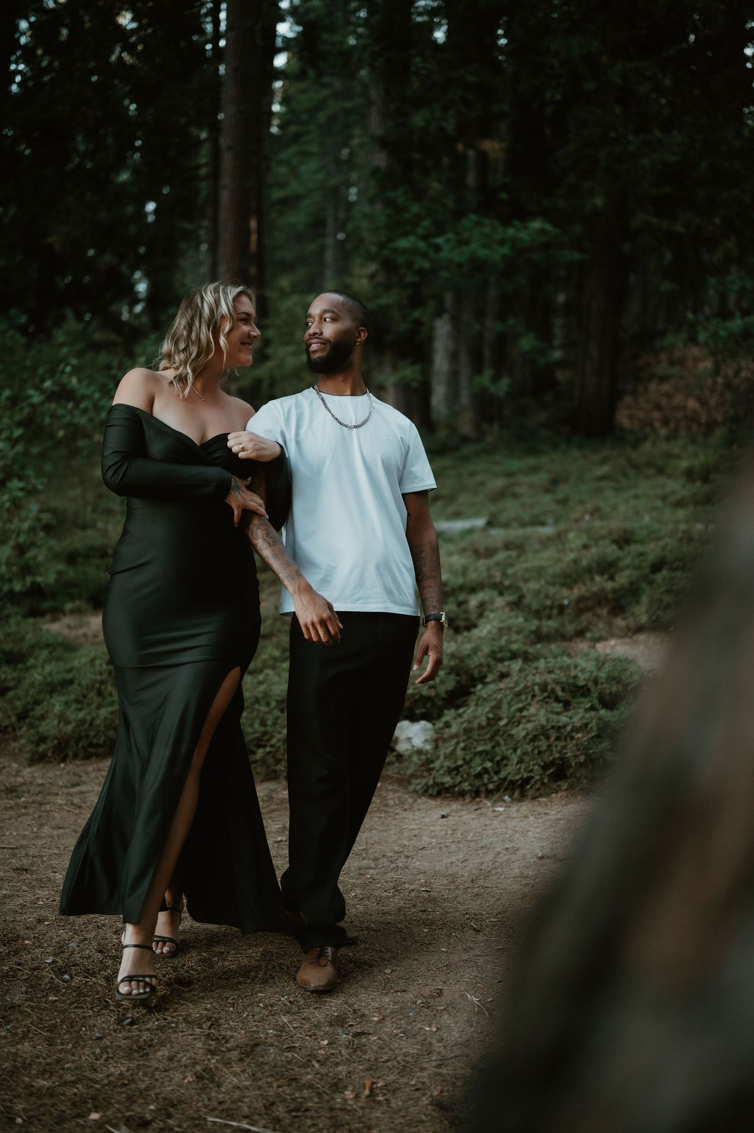 A romantic couple in contrasting black and white outfits share intimate moments while walking through a wooded forest path.