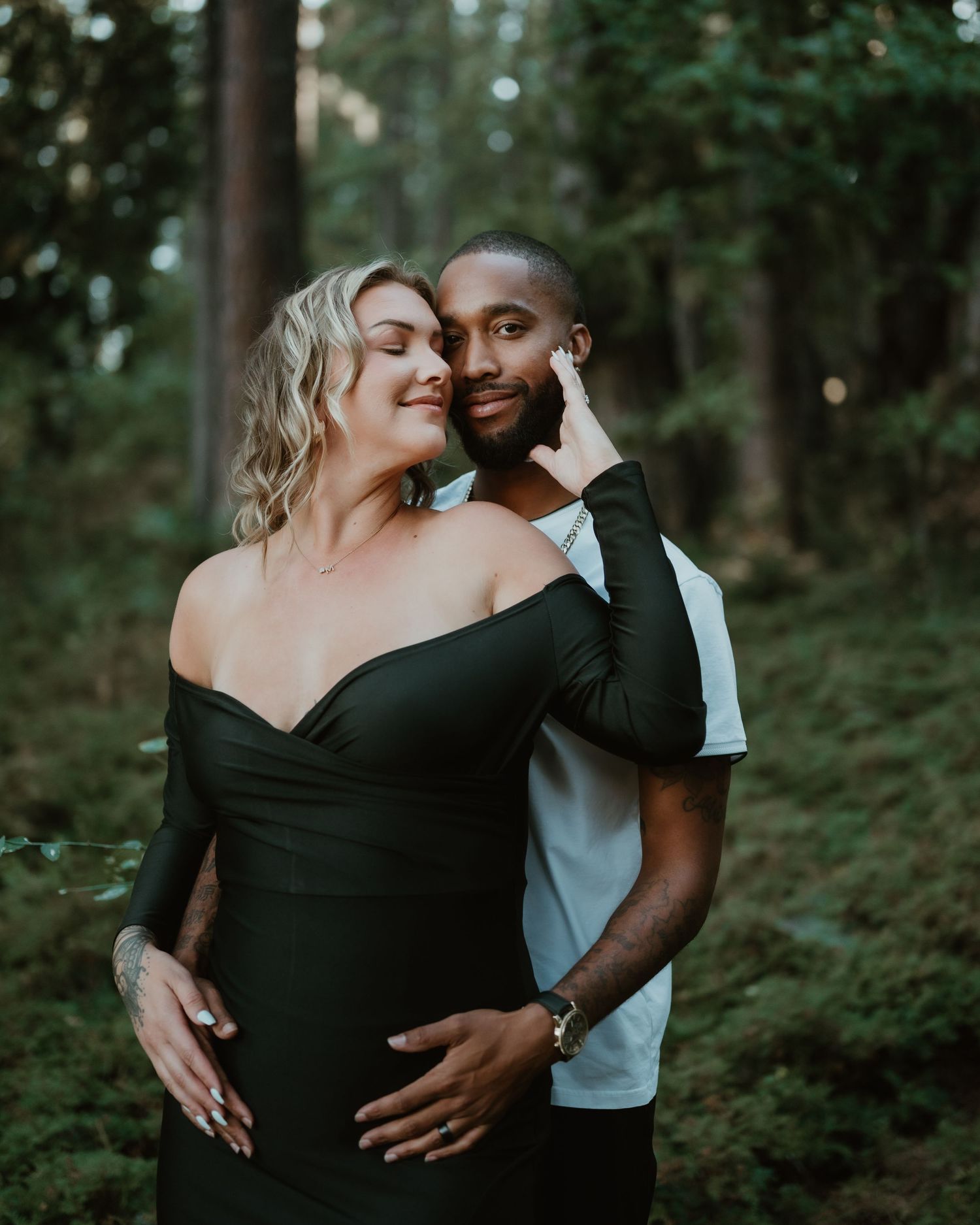 A loving couple shares an intimate moment together in a wooded setting at sunset, with her wearing a black dress.