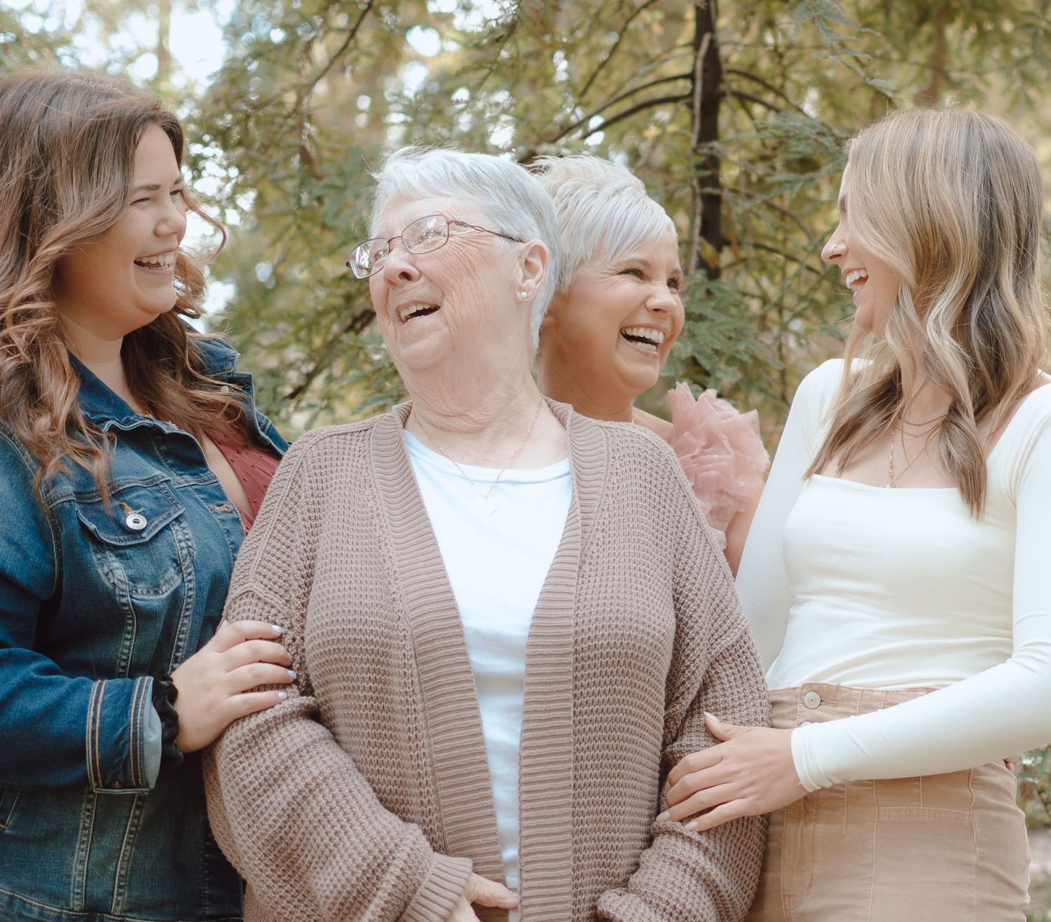 A group of women laughing and embracing while wearing cozy sweaters outdoors on a sunny day.