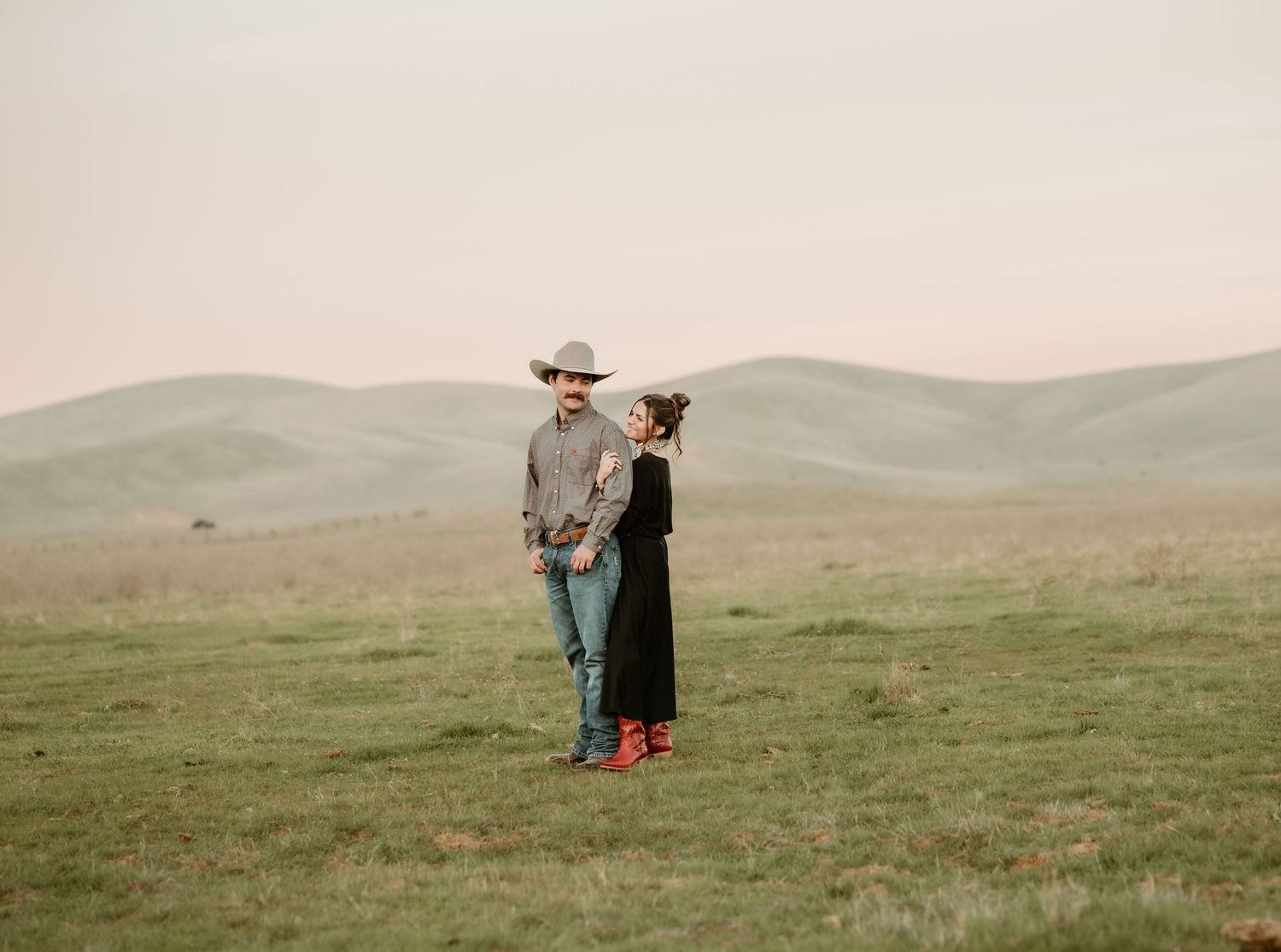 Two people stand together on a misty green hillside in matching red boots and hats.