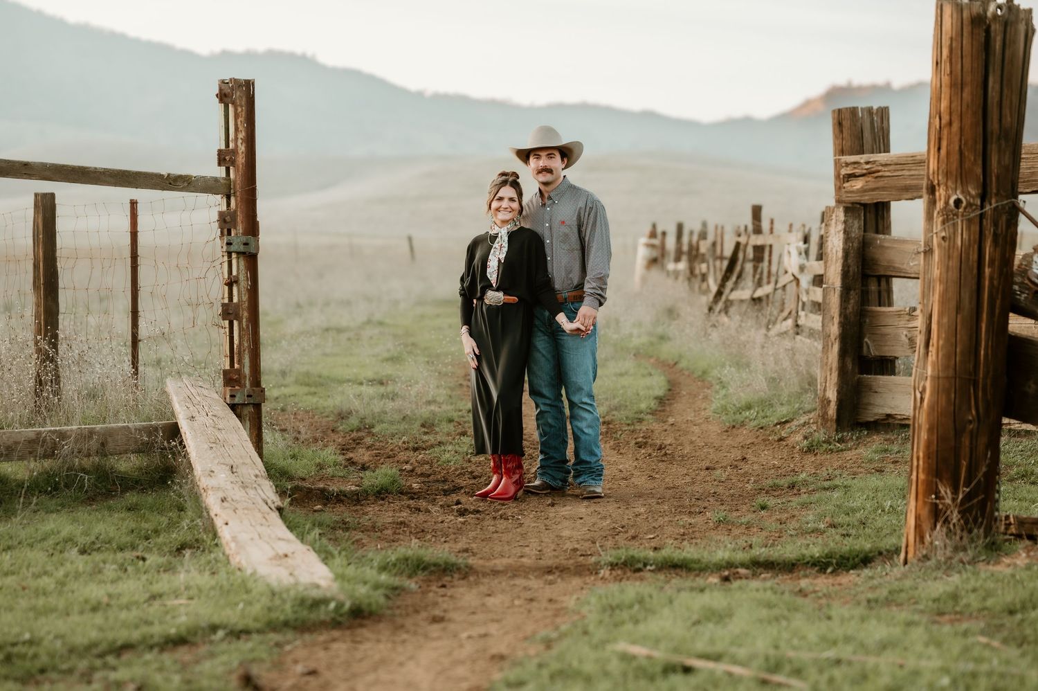 A couple stands together at a rustic wooden gate on a misty mountain ranch with rolling hills in the background.