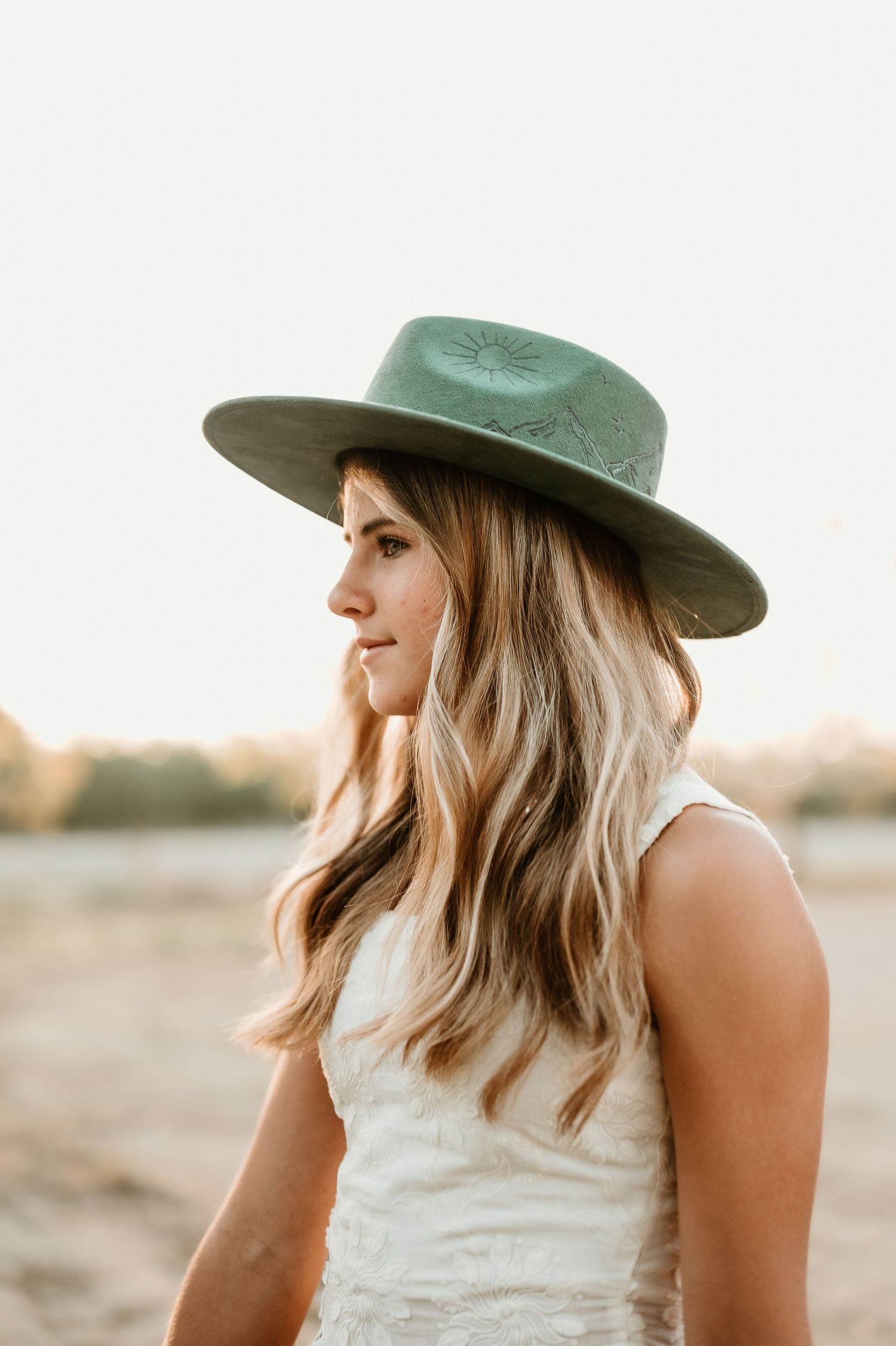 Portrait of person in a wide brimmed hat and white sleeveless top against a soft sunset backdrop.