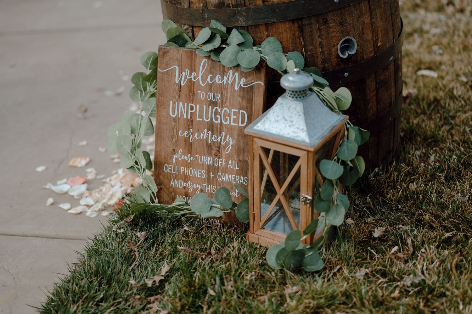 Rustic unplugged ceremony sign with lantern and eucalyptus leaves against wooden barrel.