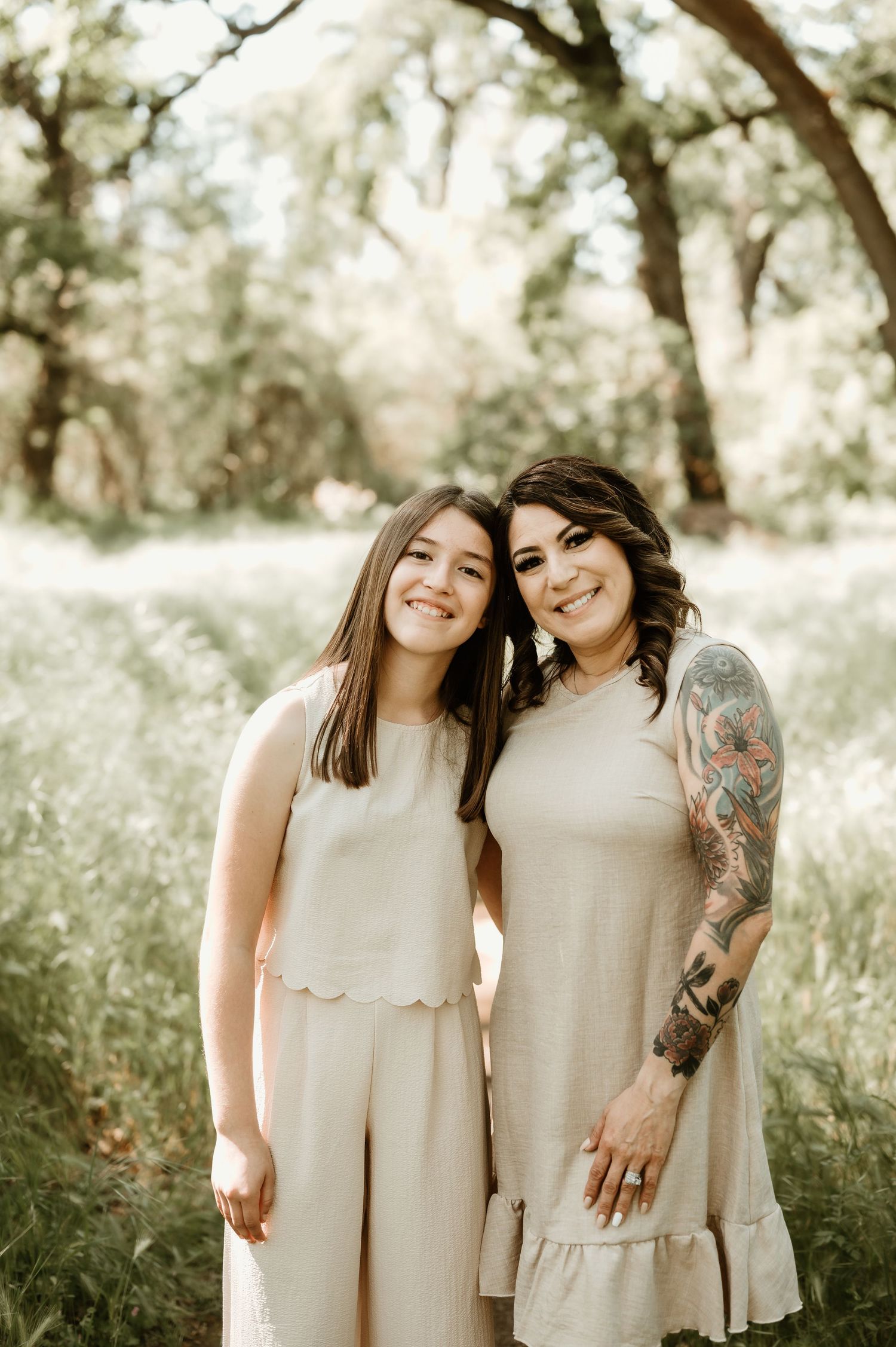 Two people in matching beige tiered dresses pose together in a sunlit outdoor setting with trees in the background.