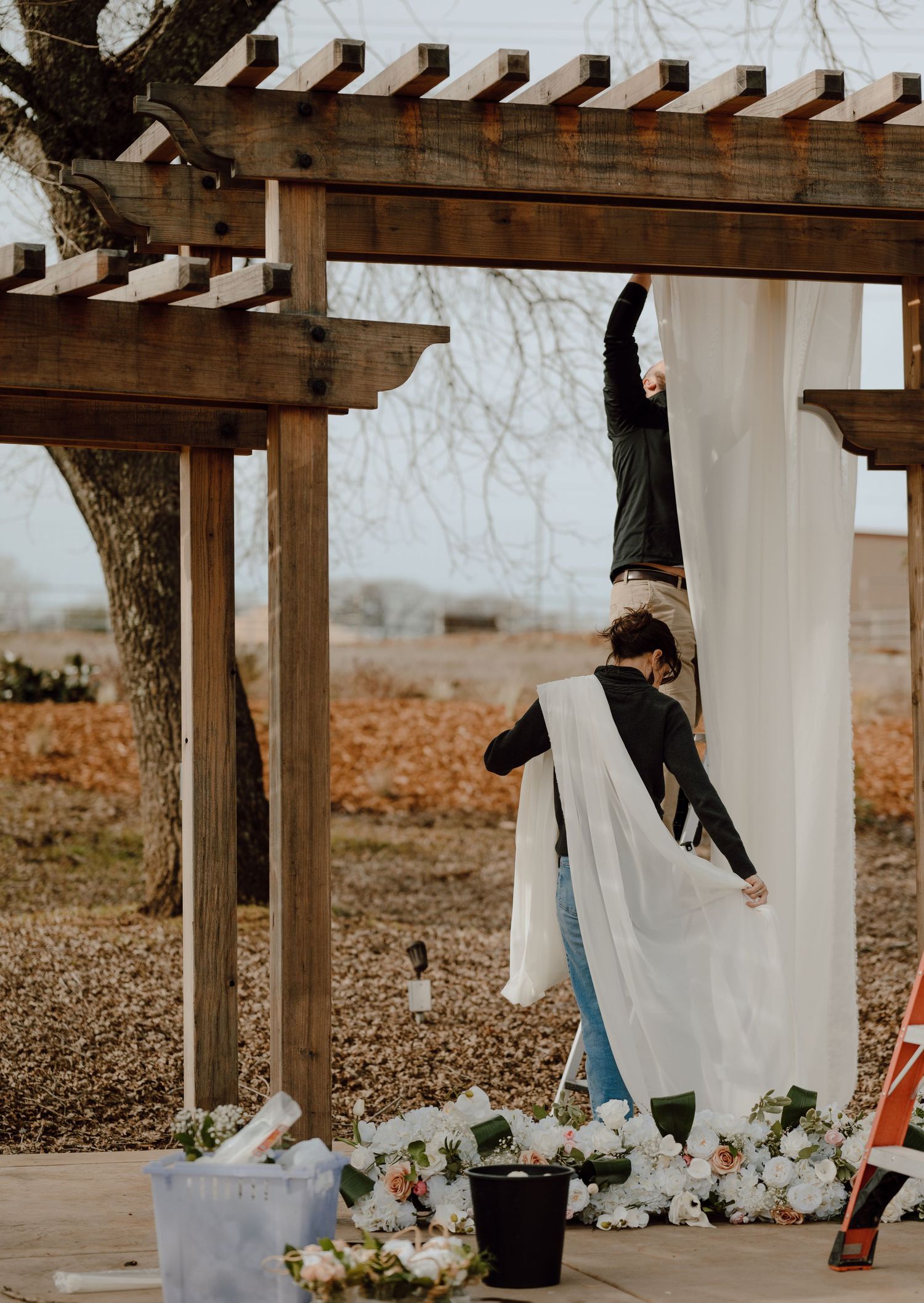 Someone hanging white fabric on a wooden pergola during event setup.