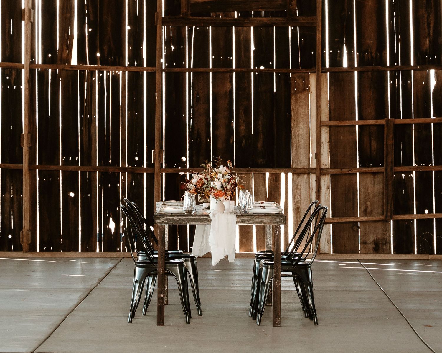 Rustic barn wedding setup with industrial chairs and floral centerpiece on wooden table.