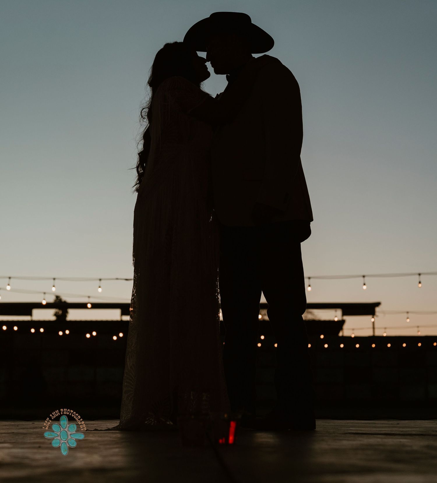 Silhouette of couple in western wear embracing at sunset with string lights in background.