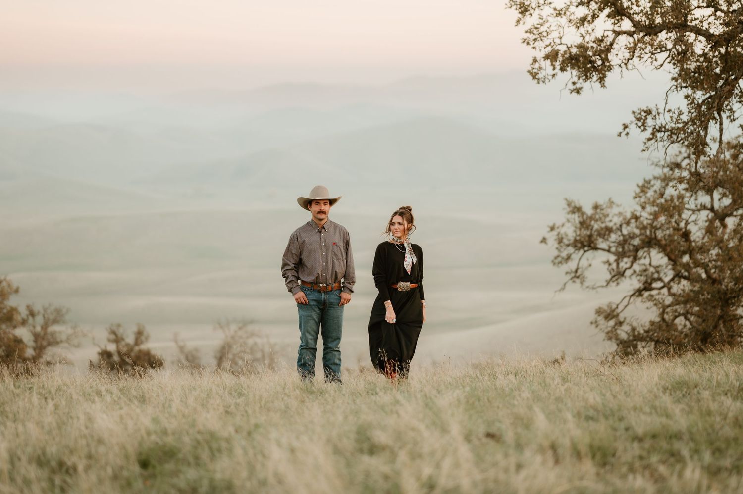 A couple walks together through a scenic meadow with mountains and clouds in the background at sunset.