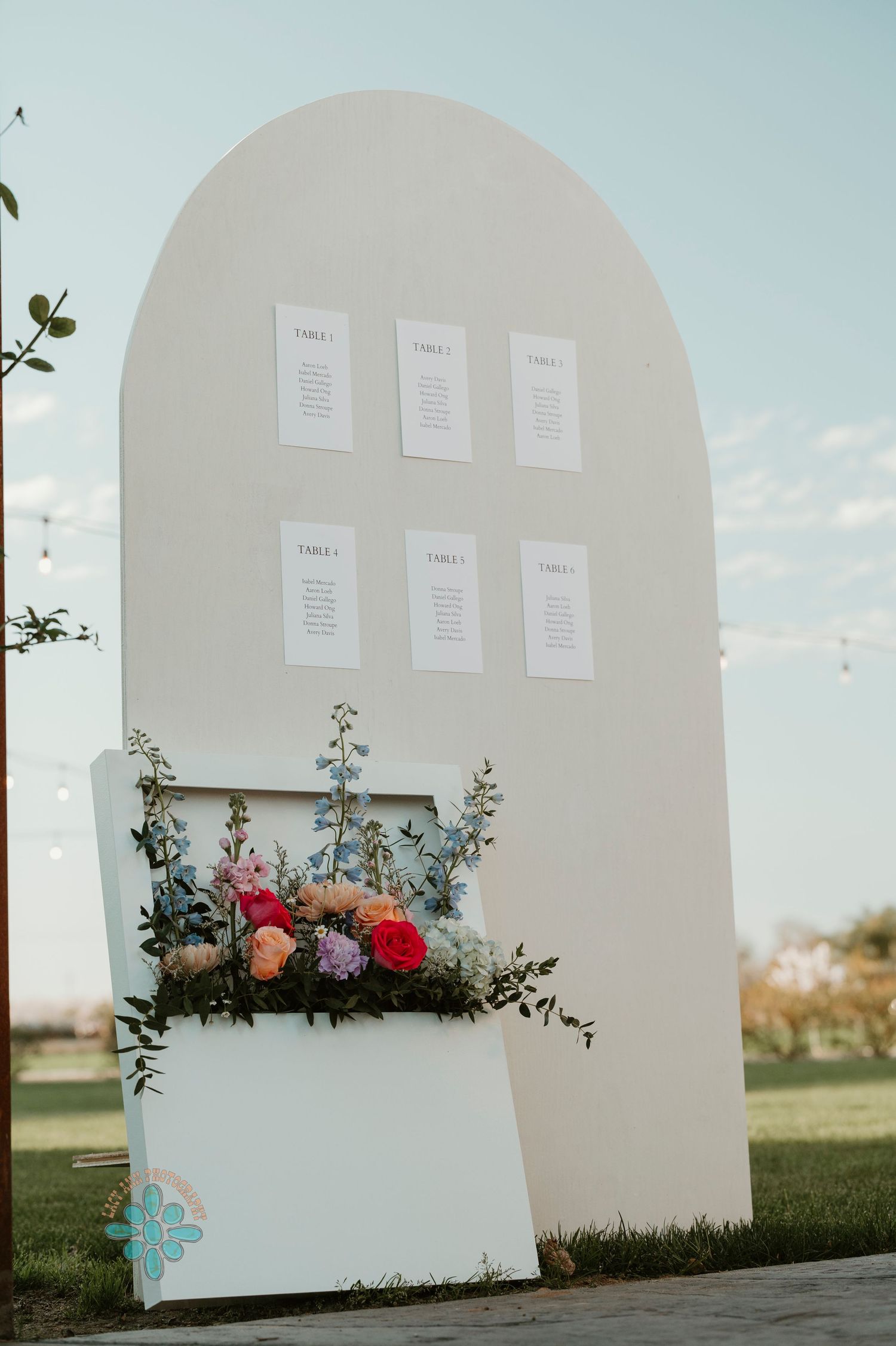 White curved archway with seating chart displays and floral arrangement at an outdoor wedding venue.